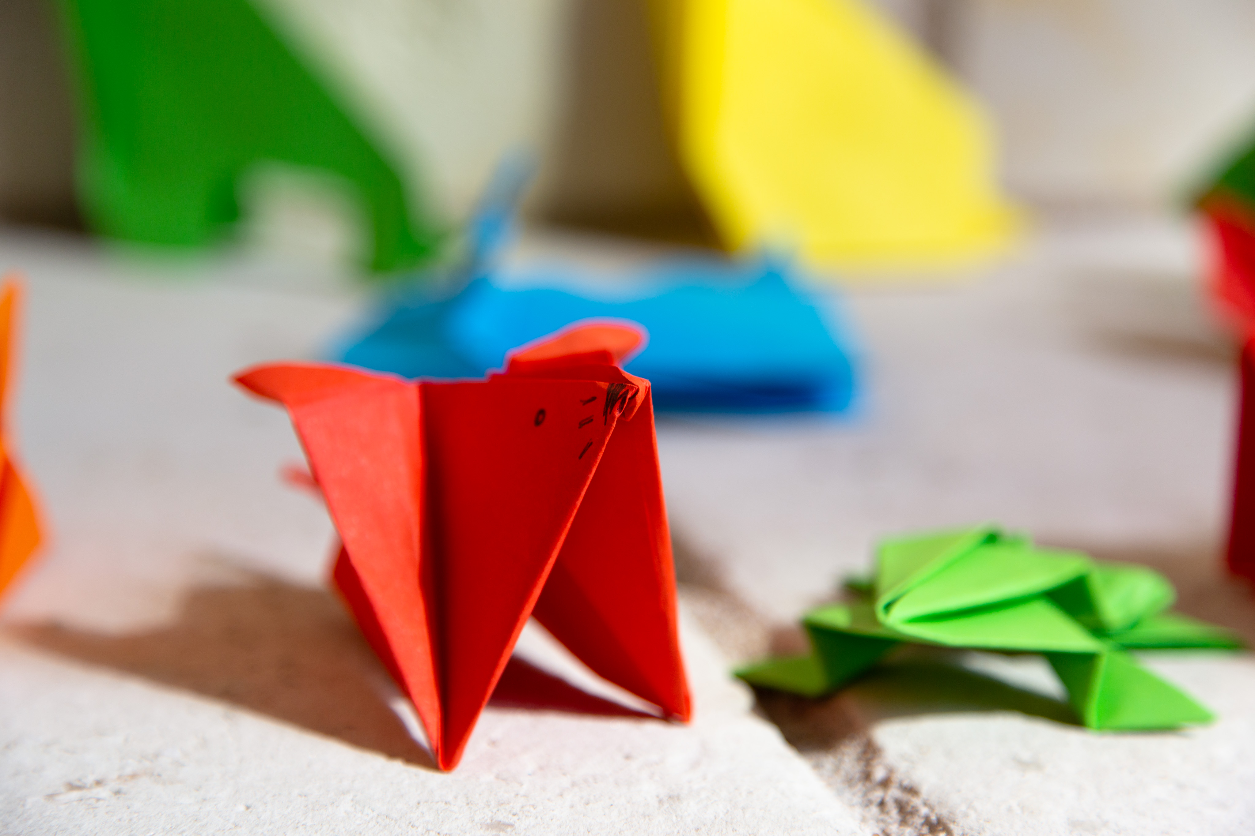Colorful origami animals sitting on a stone bench. In the center of the frame is a small red mouse origami. To the right is a flat green frog origami.