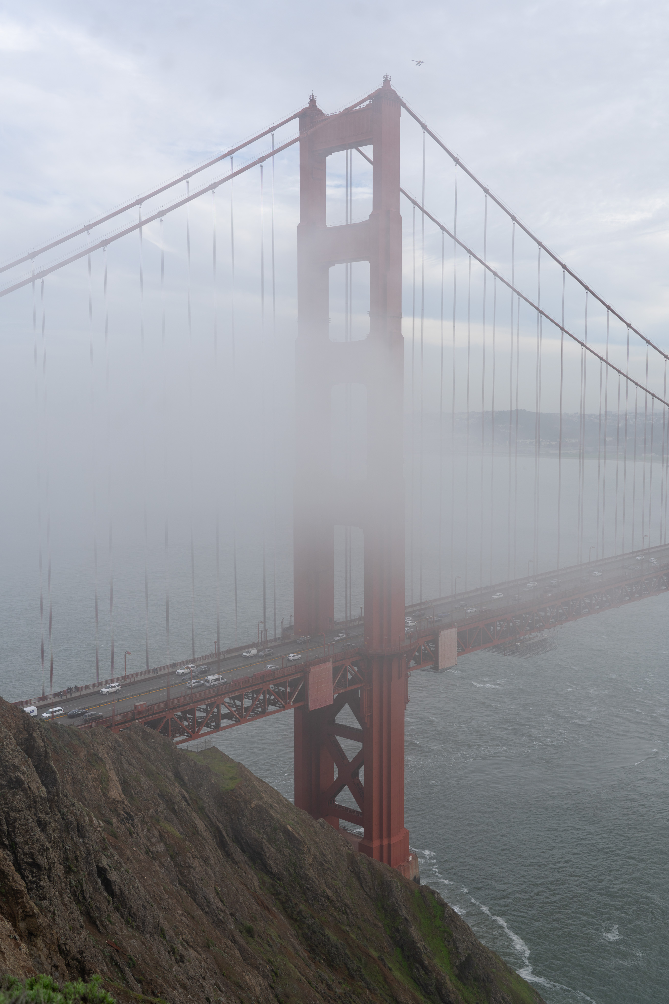 The northern tower of the Golden Gate bridge, partially obscured by fog/clouds. A small plane can be seen passing over.