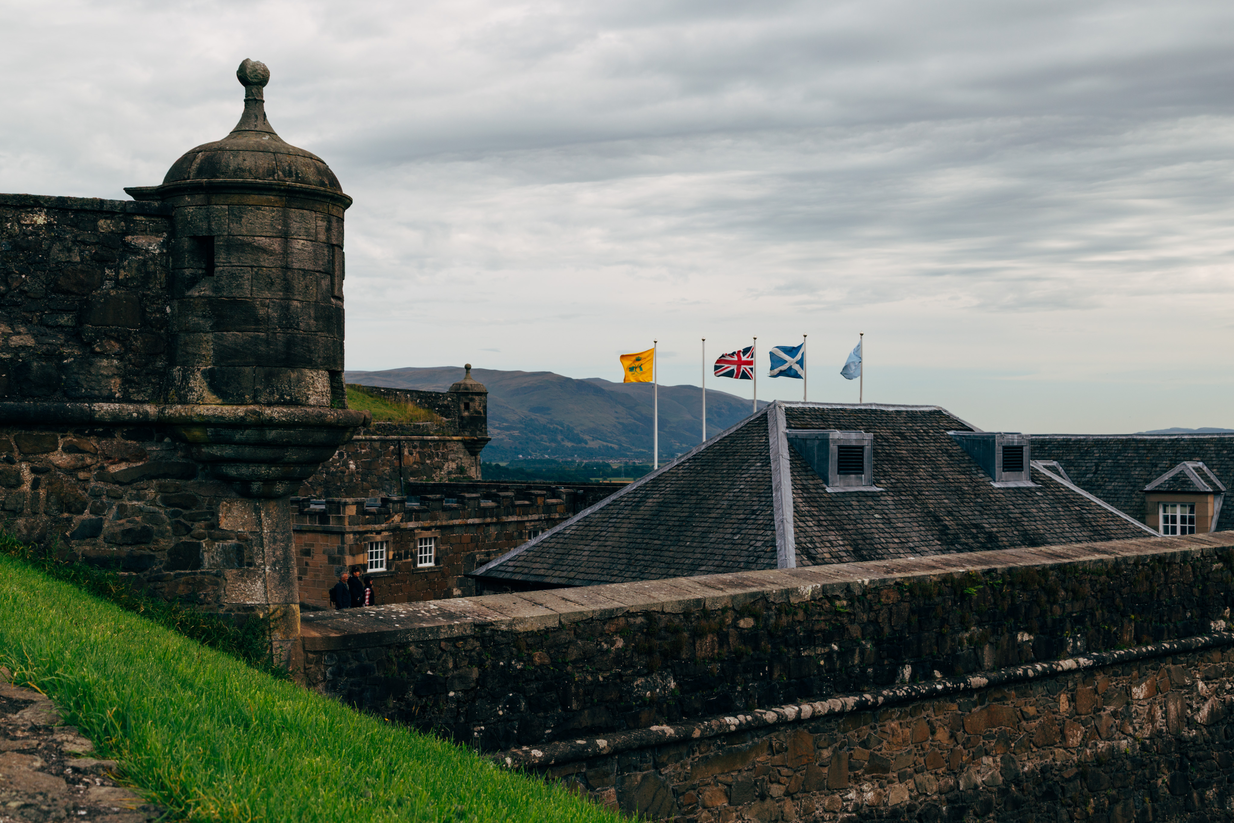 A view from buildings at Stirling castle. A corner turret is on the left of the frame — in the middle is the roof of a small building, with four flags flying. the two flags in the middle are the Union Jack of the UK and the Scottish flag .