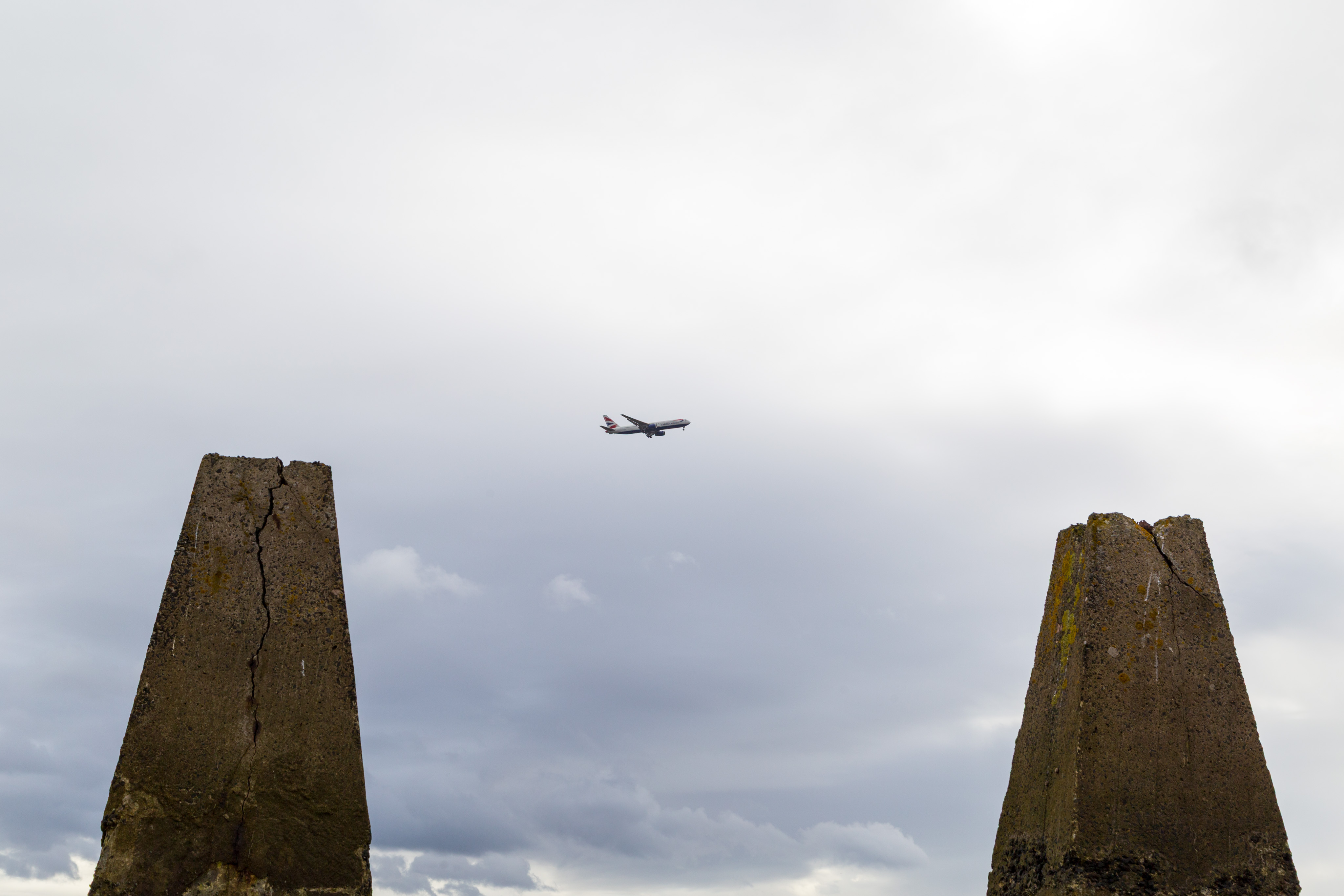 A British Airways aircraft on its landing approach to the Edinburgh airport. Two large pyramid-shaped stone pillars are in the near-ground. The sky is grey and cloudy.