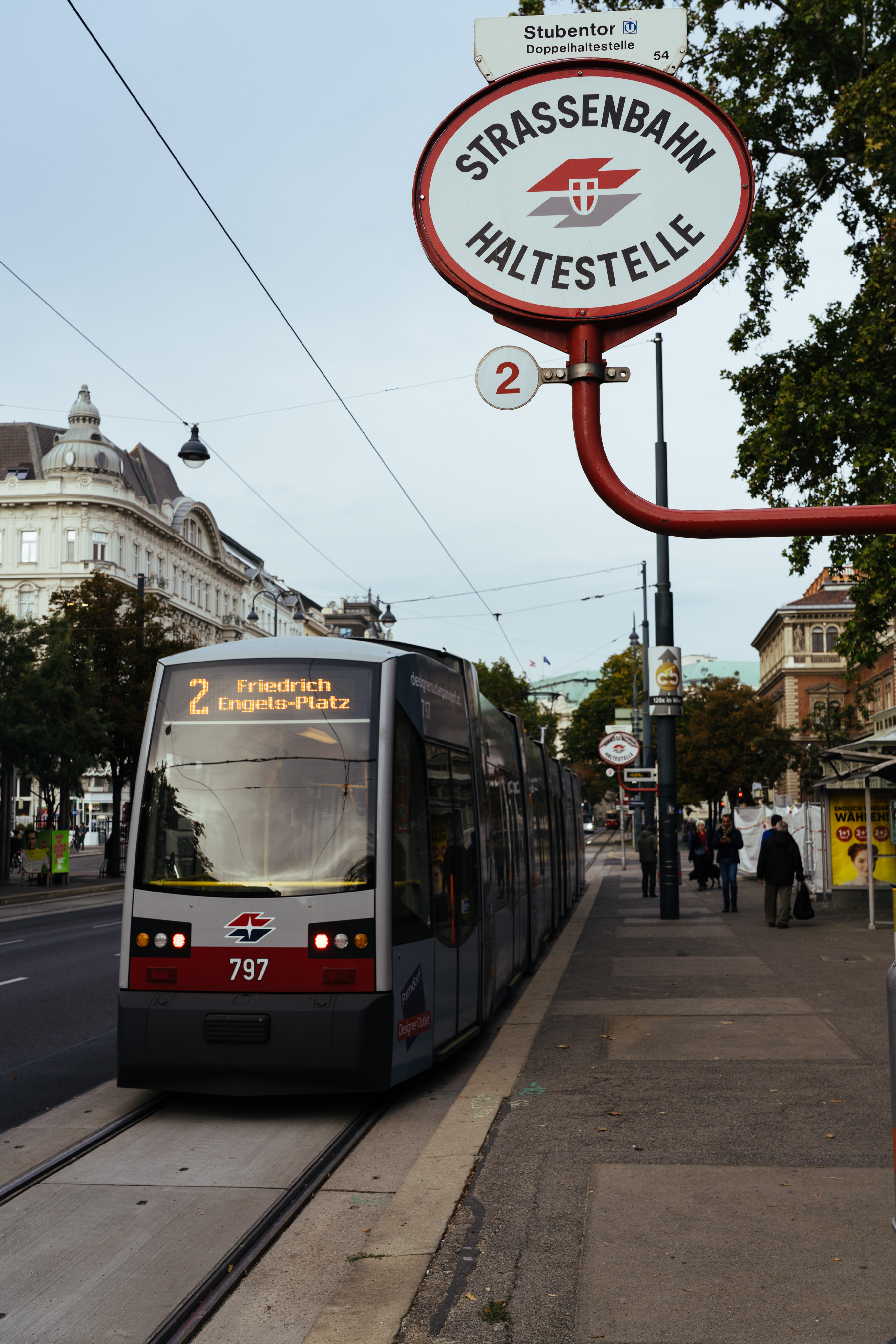 A streetcar stopped at a streetcar stop. The streetcar has a destination of Friedrich Engels-Platz. The streetcar stop sign is just visible in the upper right of the frame, reading "Strassenbahn haltestelle"