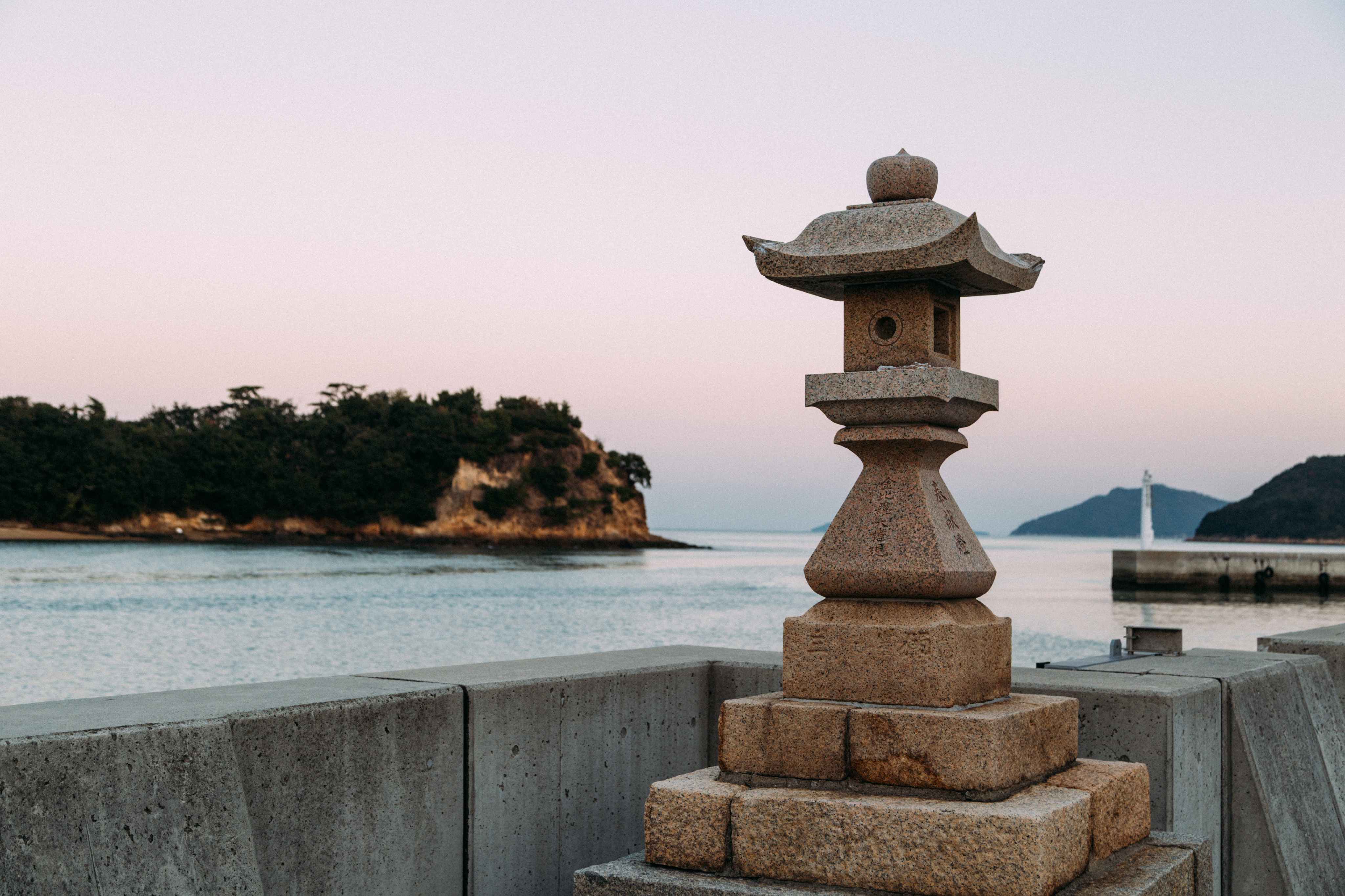 A lantern in the foreground with a small harbor in the background. The harbor leads out into the Japanese inland sea, with a rocky and tree-covered piece of land in the background.