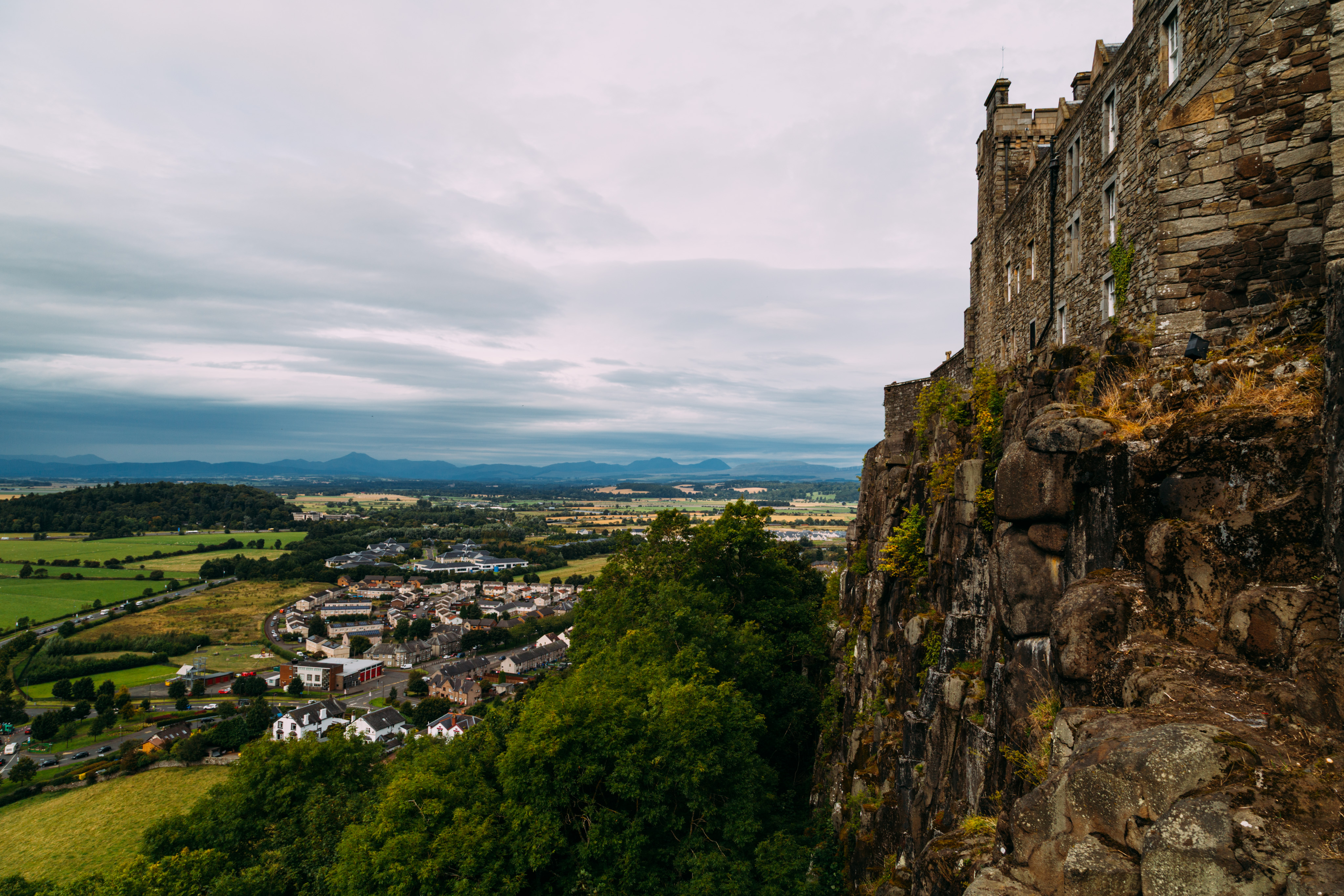 A view from above the town near Stirling castle. A number of shops and homes can be seen in patterns. To the left and behind the town is Scottish countryside and some forests. On the right side of the frame is a cliff atop which buildings of Stirling castle sit.