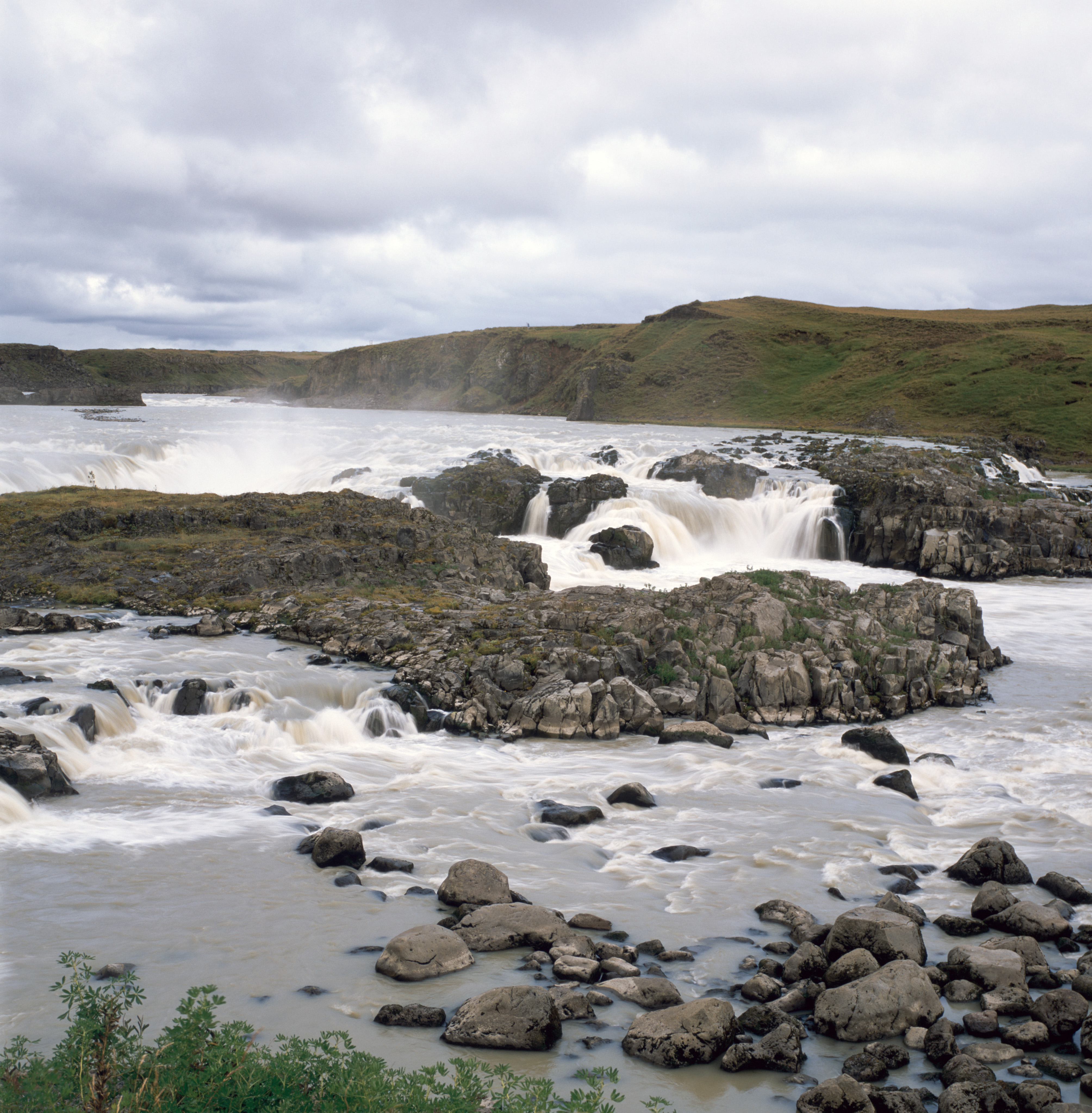 A small, wide waterfall flows over rocks. There are green hills and cliffs in the background and black rocks studded throughout the stream of water. The water has been blurred in its flowing.