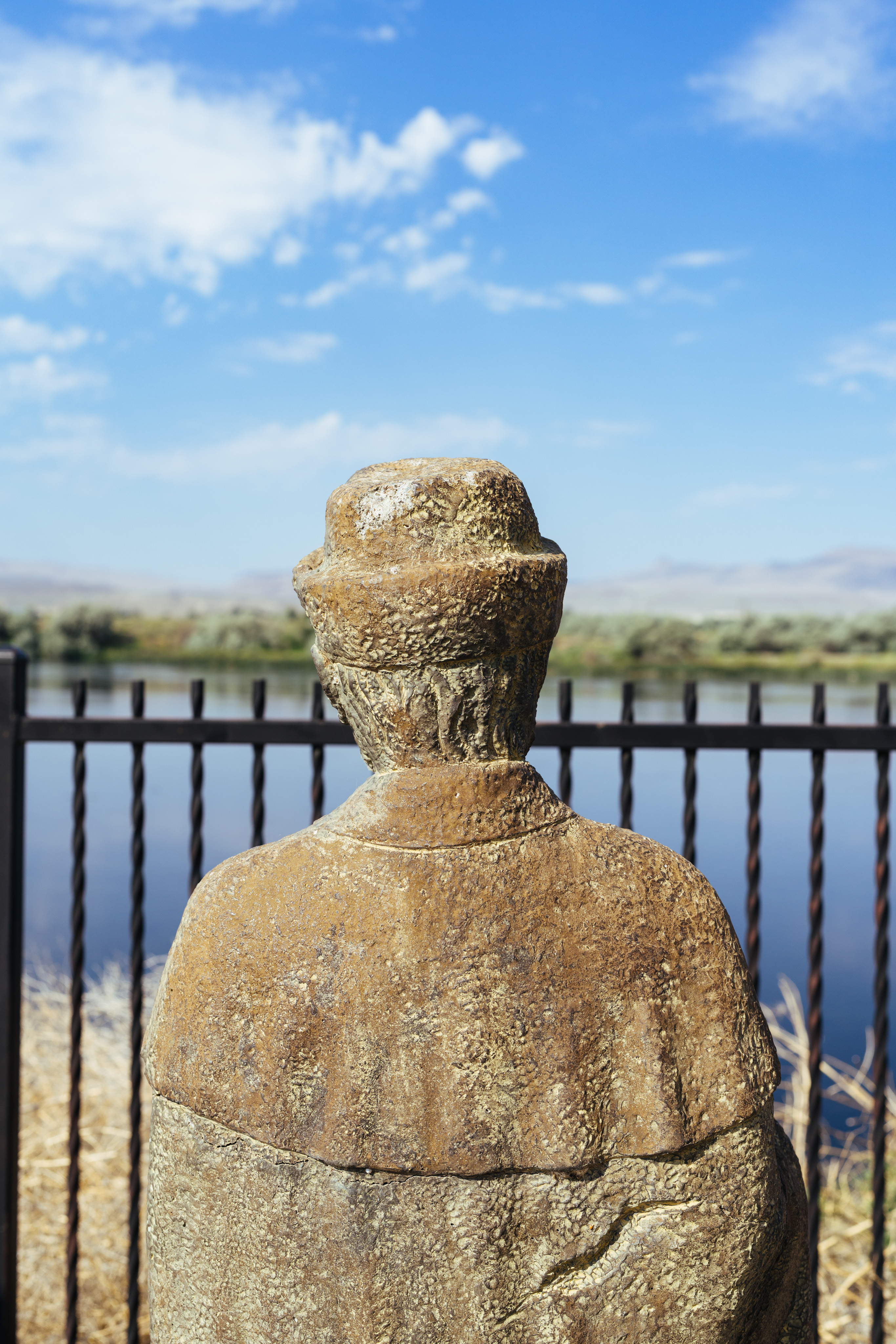 A photograph of the back of a statue of a person. The statue sits in front of (and is looking across) the Snake river. There is a black metal fence between the statue and the river and there are a few clouds in a blue sky