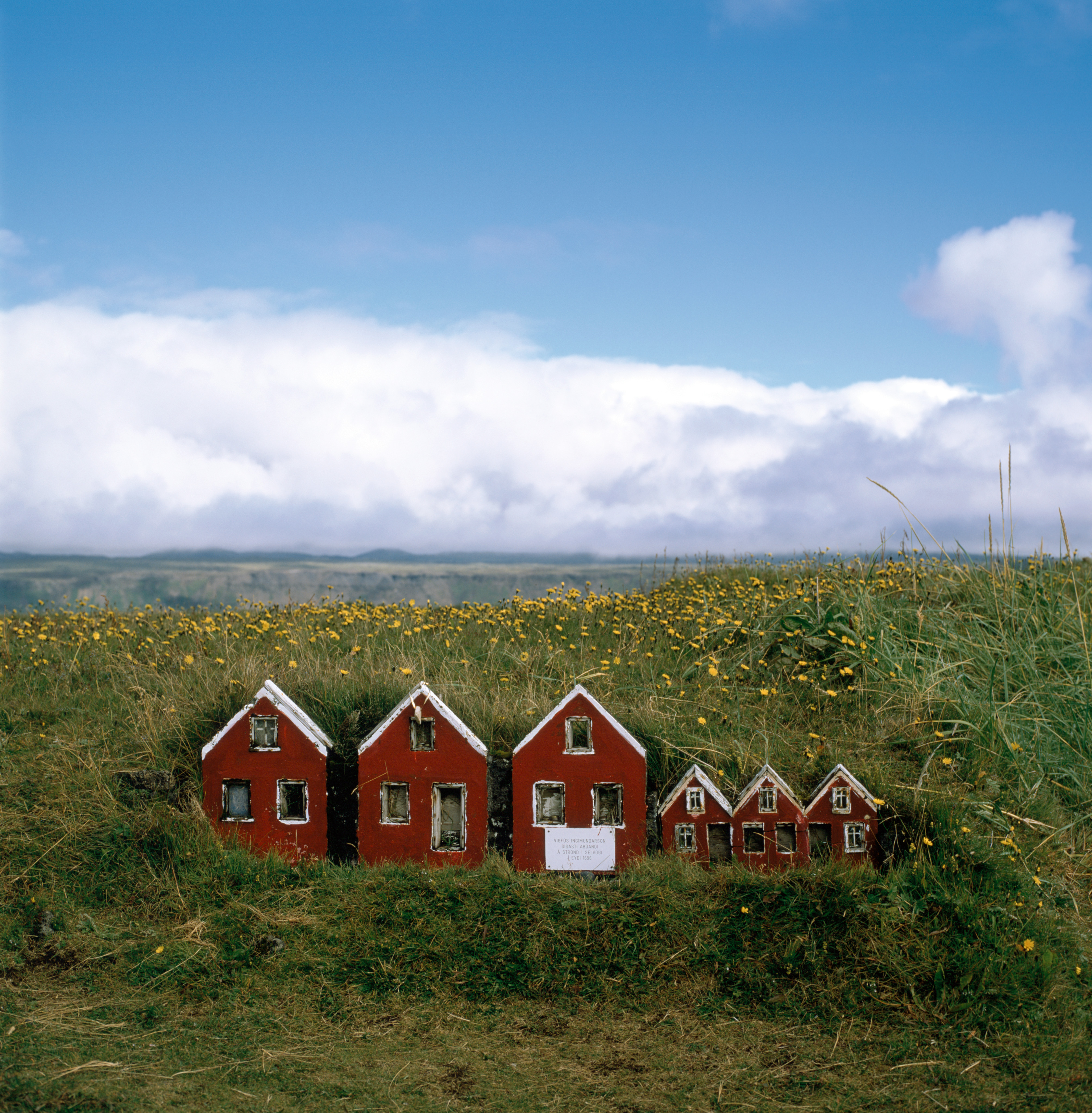 Three small models of the fronts of houses next to three even smaller models of houses stand in a meadow. Behind the houses is a field of green and yellow flowers. The sky is blue with a large mass of clouds low.