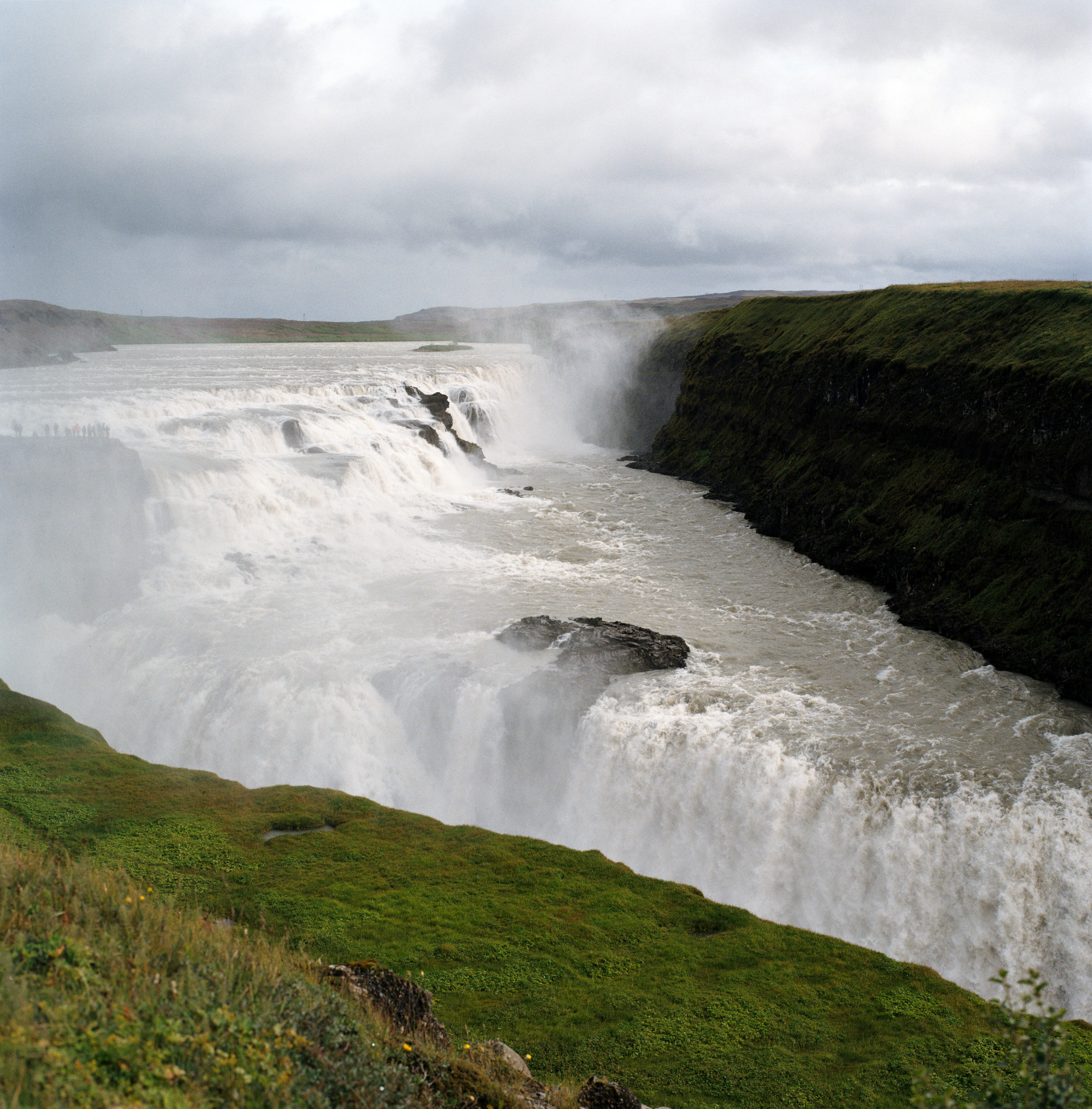 A close up of Gullfoss in Iceland. There’s a spot of green in the foreground, and the waterfalls just break above that patch. In the background, on the right are green cliffs.