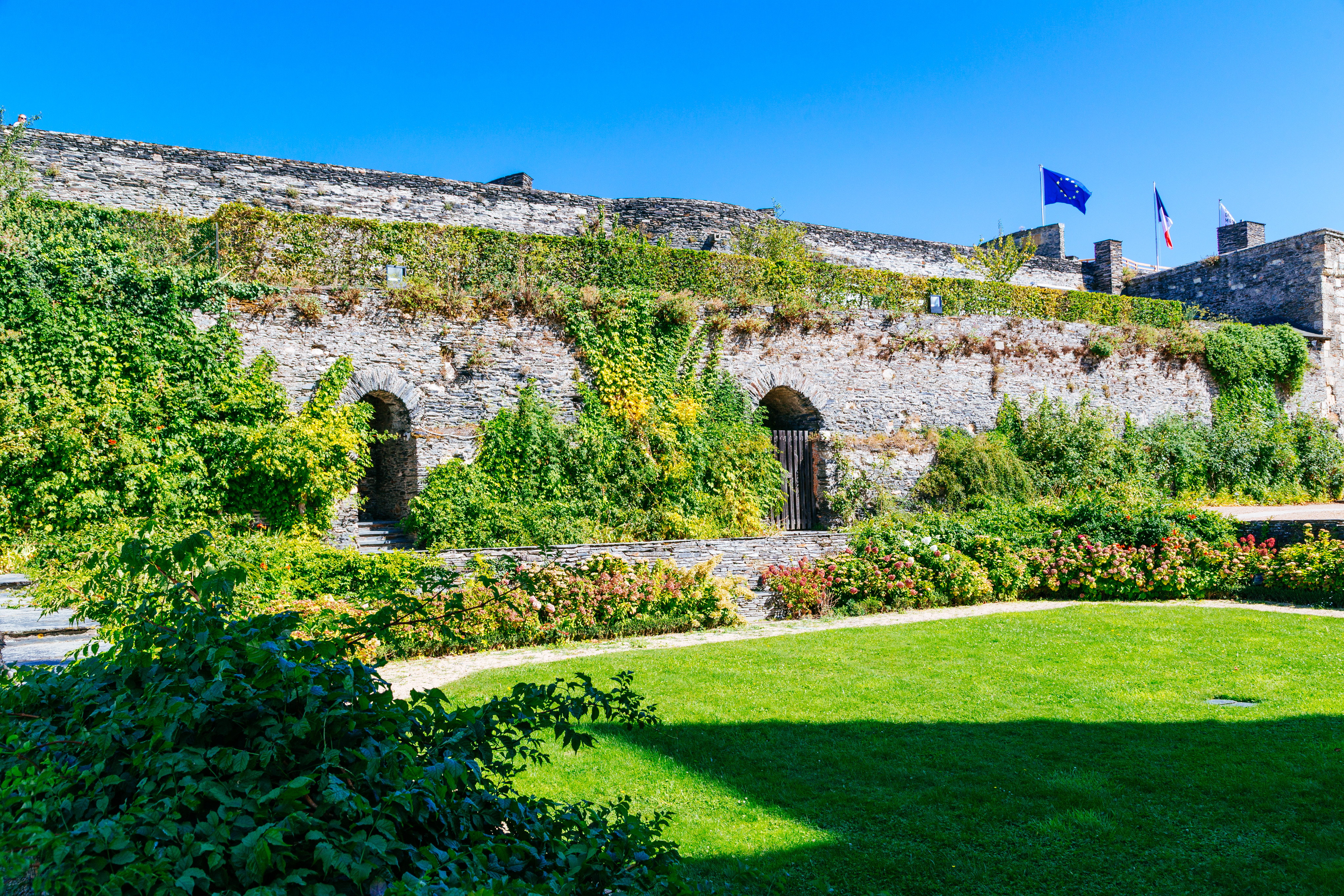 A lush garden with many vines growing on a stone wall with two doorways. In the far left of the image a flag of the EU and France can be seen.