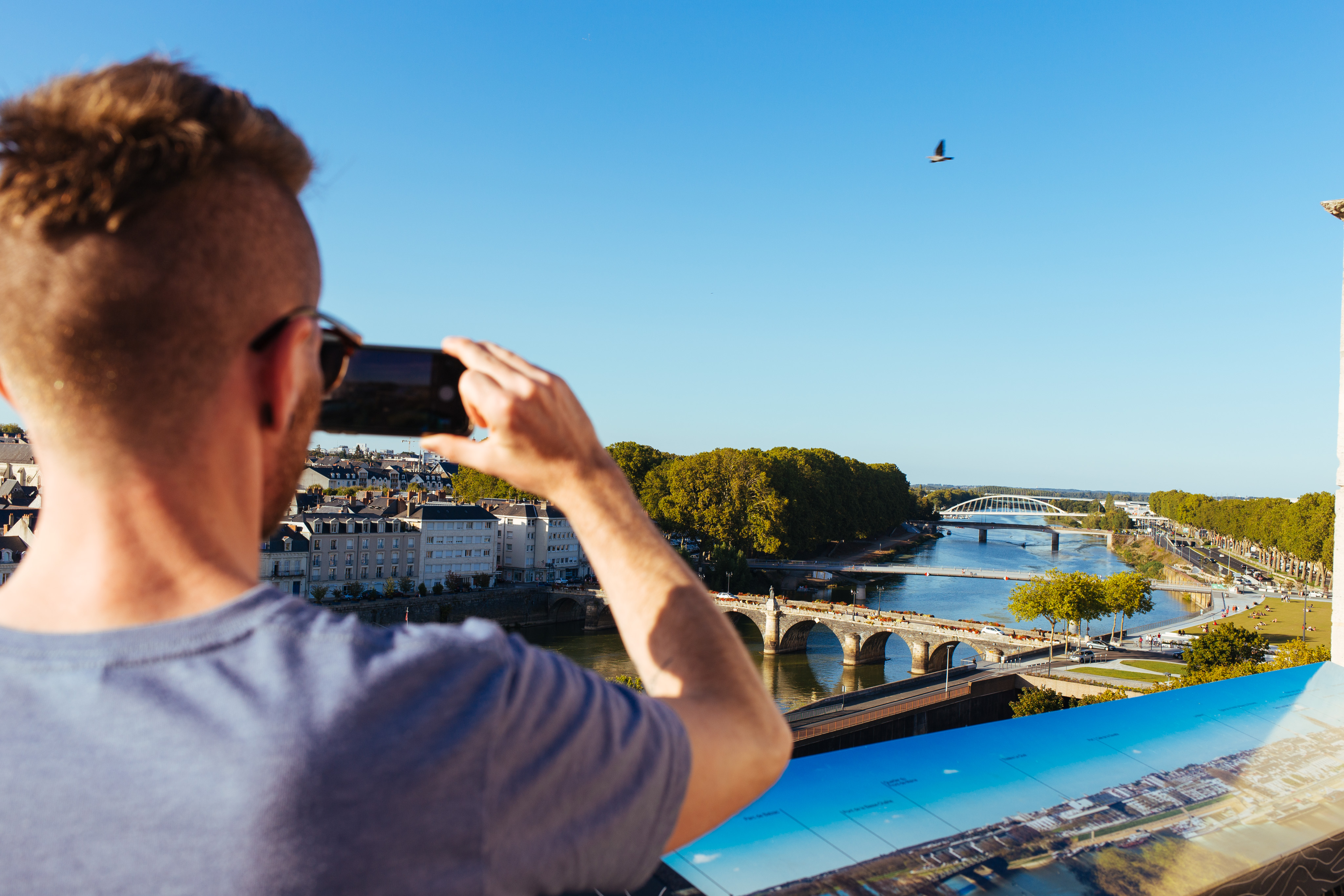 A view up the Sarthe river in Angers. Five bridges of varying construction methods can be seen crossing the river. In the foreground a person is using a phone to take the a photograph.