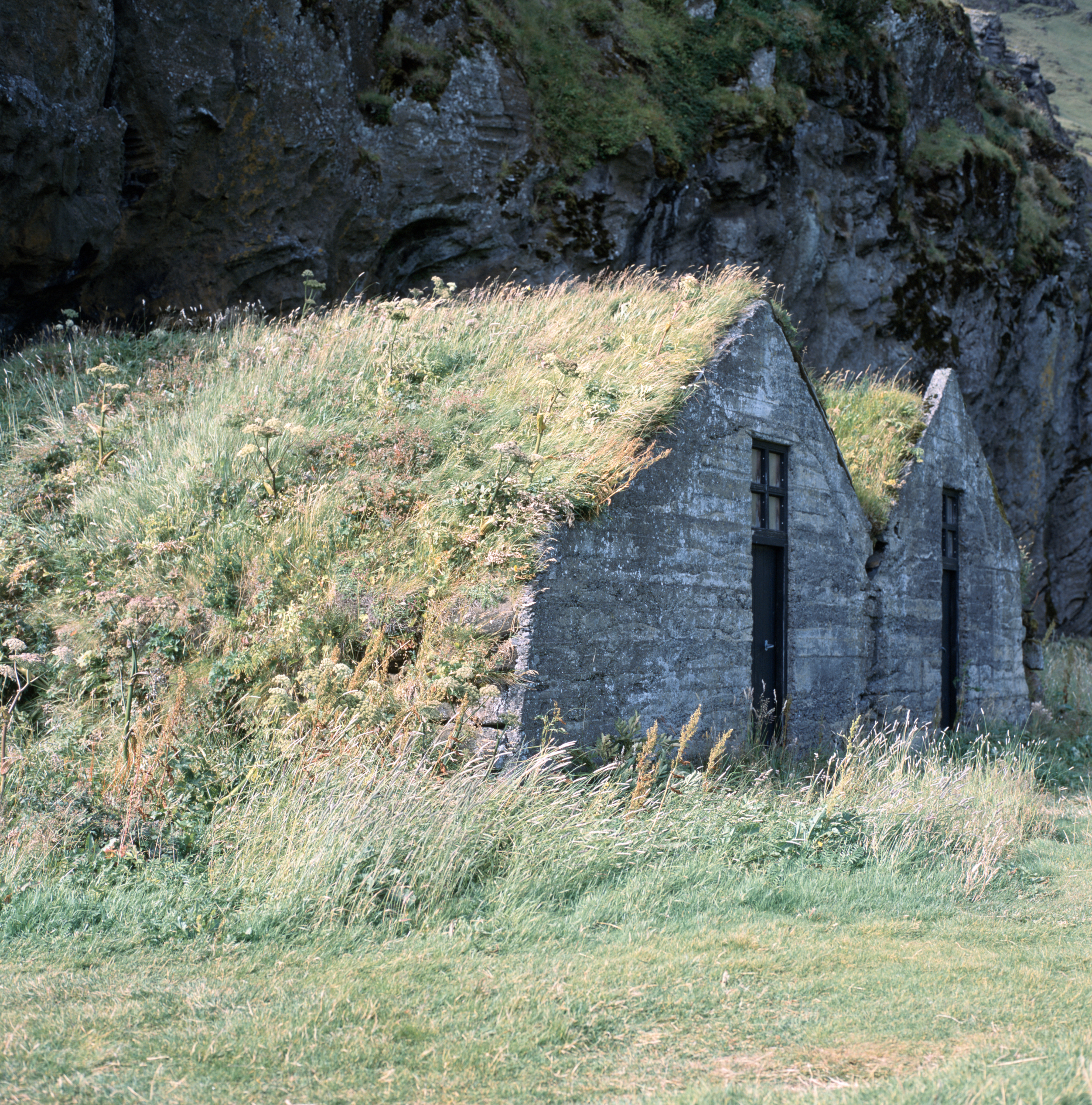 Two small houses with earthen roofs and stone fronts stand at the base of a cliff. The houses themselves are build into the cliff, and grass grows from the ground all the way up the slope of the roofs.