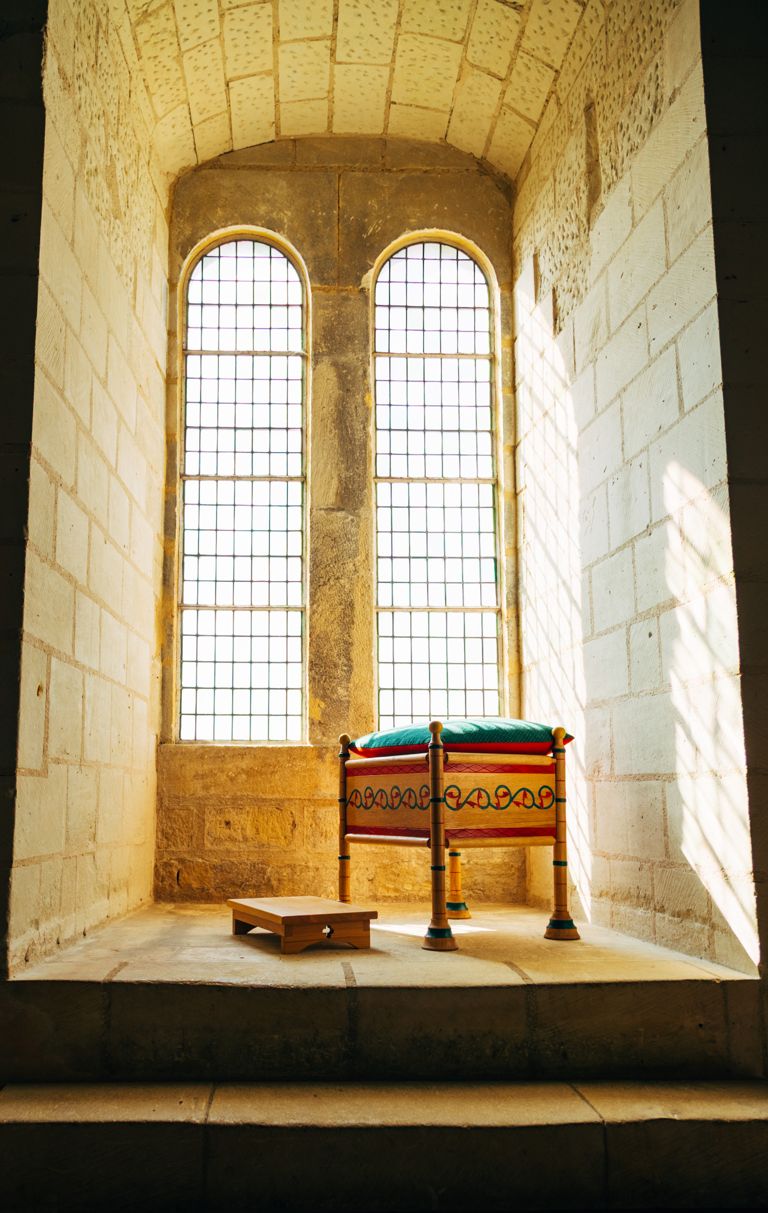 a small stool and a platform to kneel and pray on sits in a sun-drenched window inside of a castle. The stool is wood with red and green paint. The windows are two large arches with a rhythmic pattern of reinforcements / panes of glass.