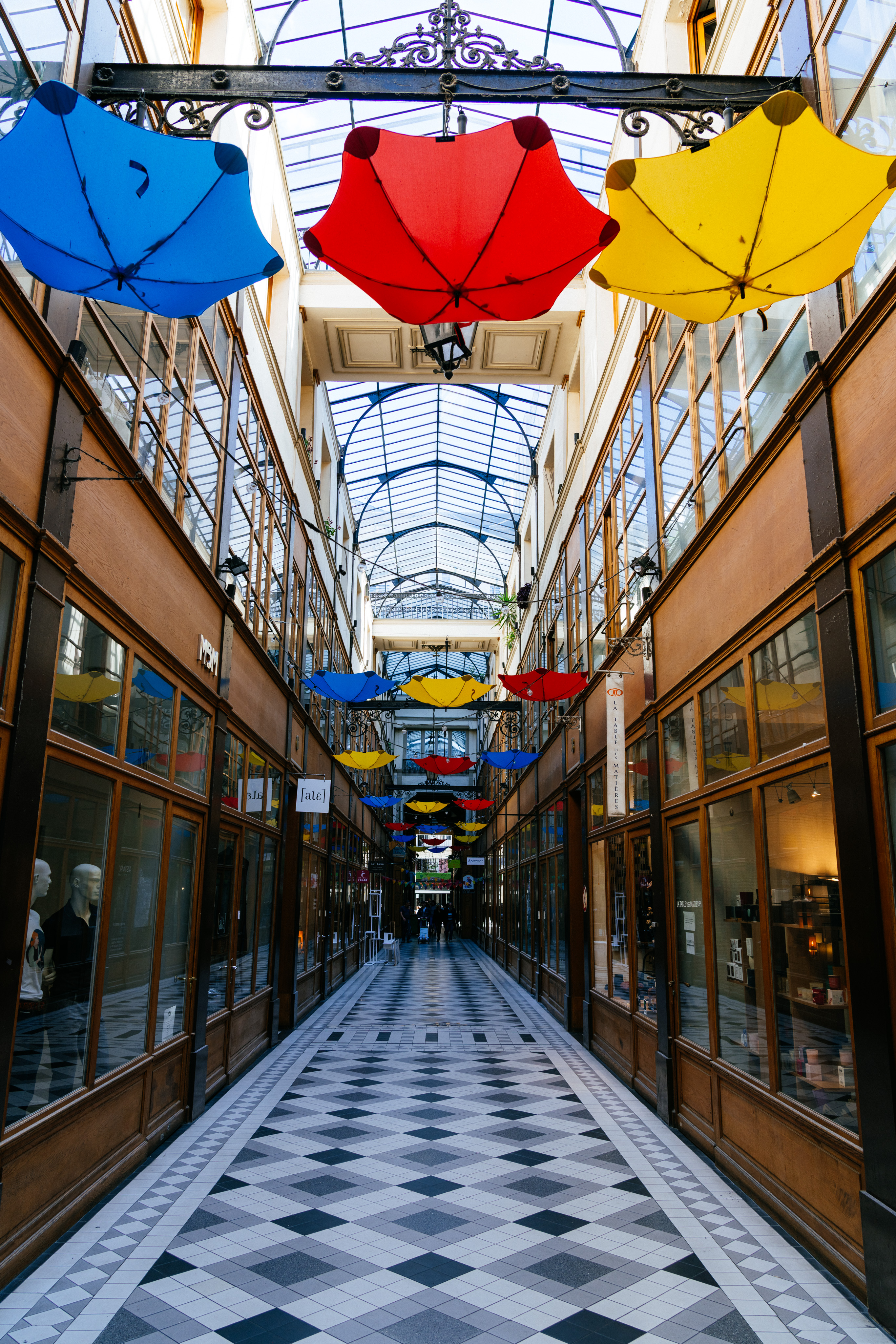 An image looking down a Parisian arcade. The floor has geometric white, black, and grey tiles. The store fronts on either side are wood and glass. Just below the glass skylights hang blue, red, and yellow umbrellas ad regular intervals down the arcade.