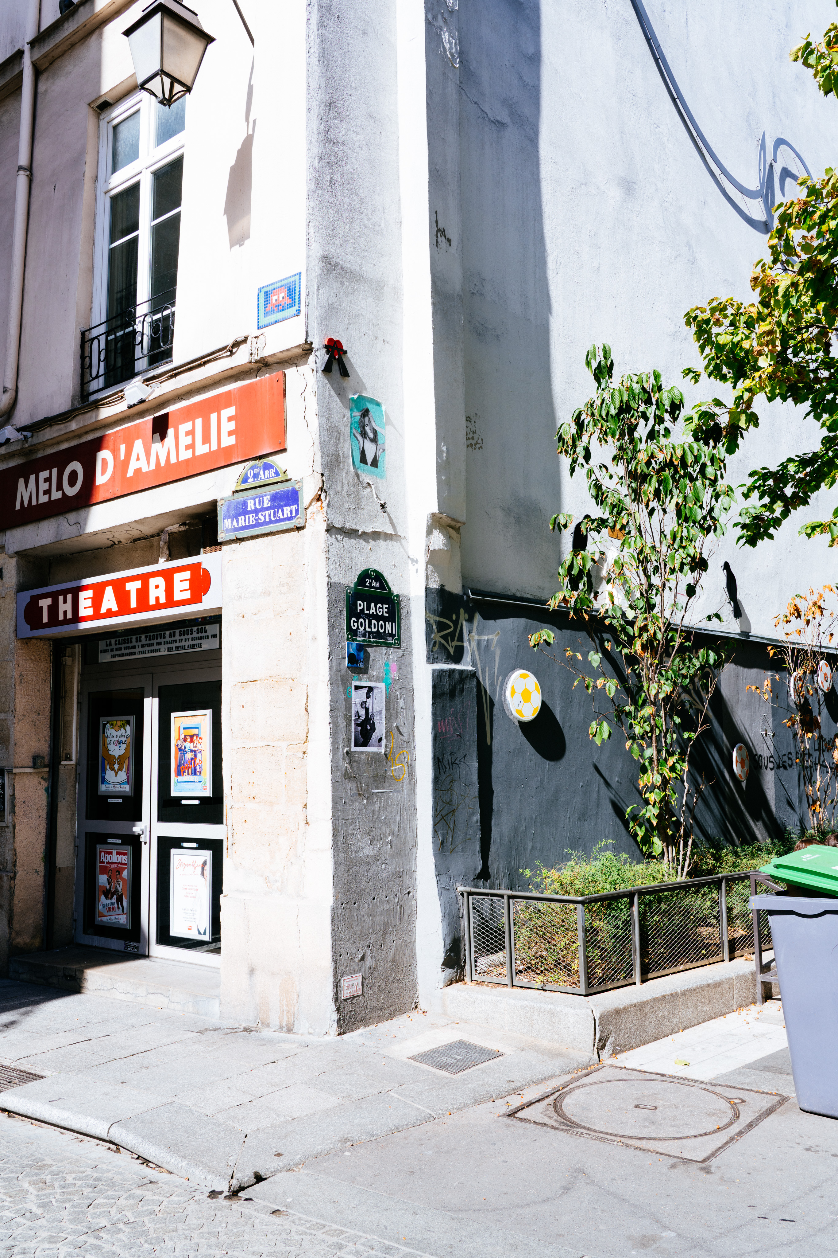 The corner of a street in Paris. The building has signs showing that it is at the corner of Rue Marie-Stuart and Plage Gordon. The building itself is white stone, with plants growing on the side. There is a large sign “Melo d’Amelie” and below that it says “theatre”. There are a number of posters on the door and the side of the building.