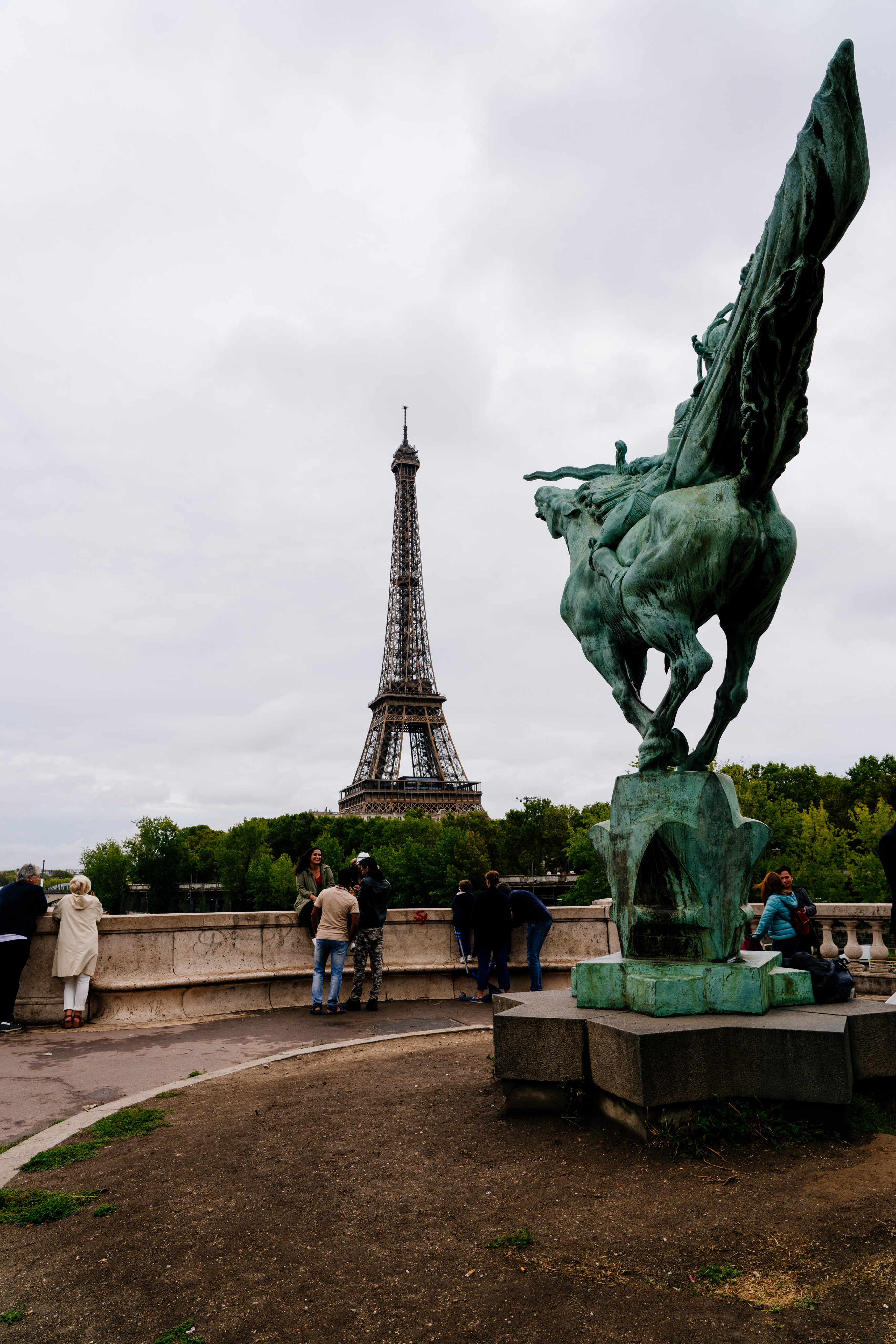 A large copper statue of a warrior on a horse is on the right side of the frame. The warrior is holding a sword that appears to be pointed directly at the Eiffel tower, which stands in the background. A number of people hangout on benches lining the area around the statue.