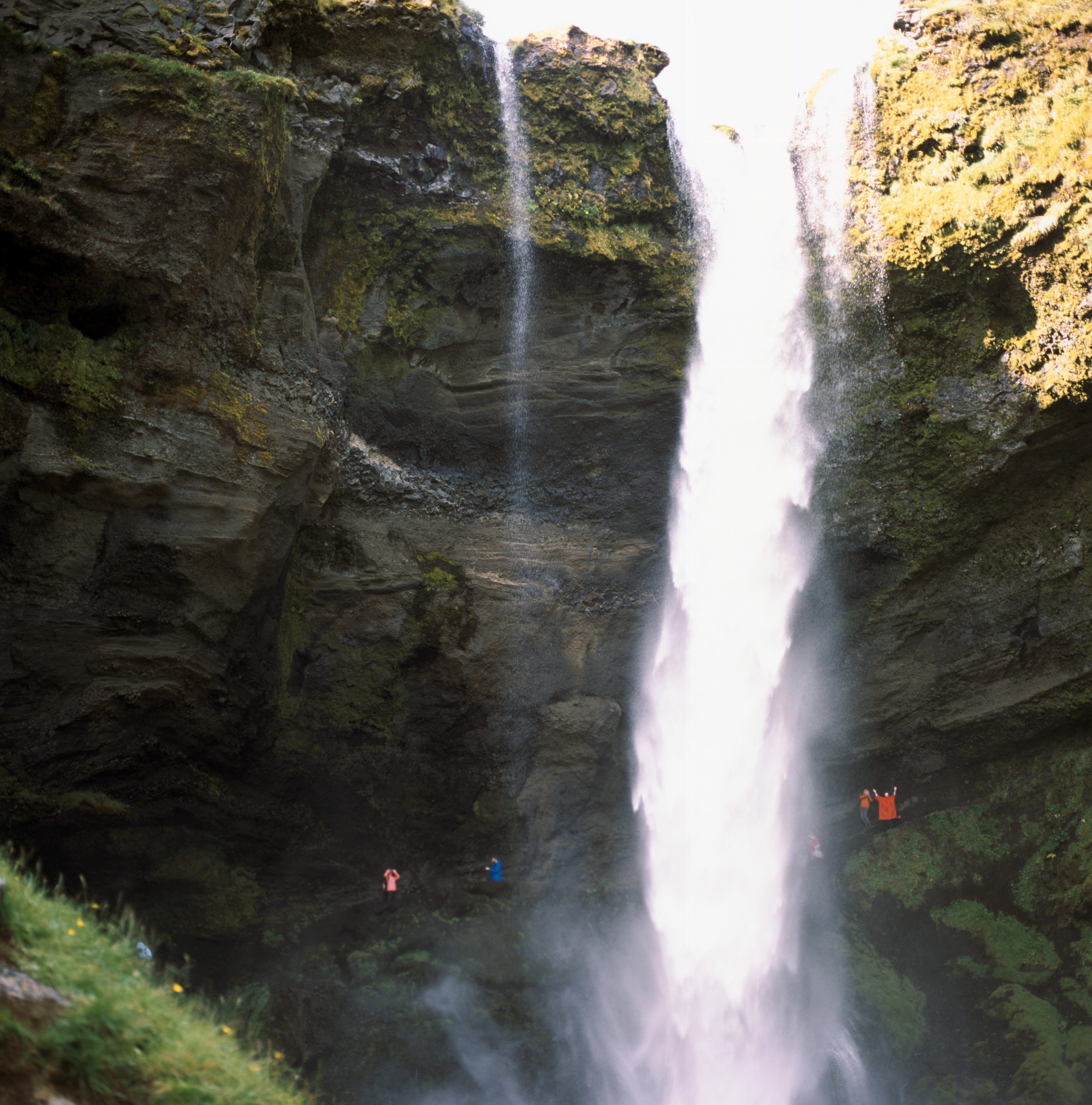 Inside a cavern containing a waterfall. The frame is almost entirely filled with cliff faces inside of the valley. There is a skinny waterfall that flows from the top to the bottom of the frame. The cliffs are rugged and craggy. There are a number of people in colorful ponchos behind the waterfall. One of the figures, in a red poncho has their hands raised (seemingly) in triumph.