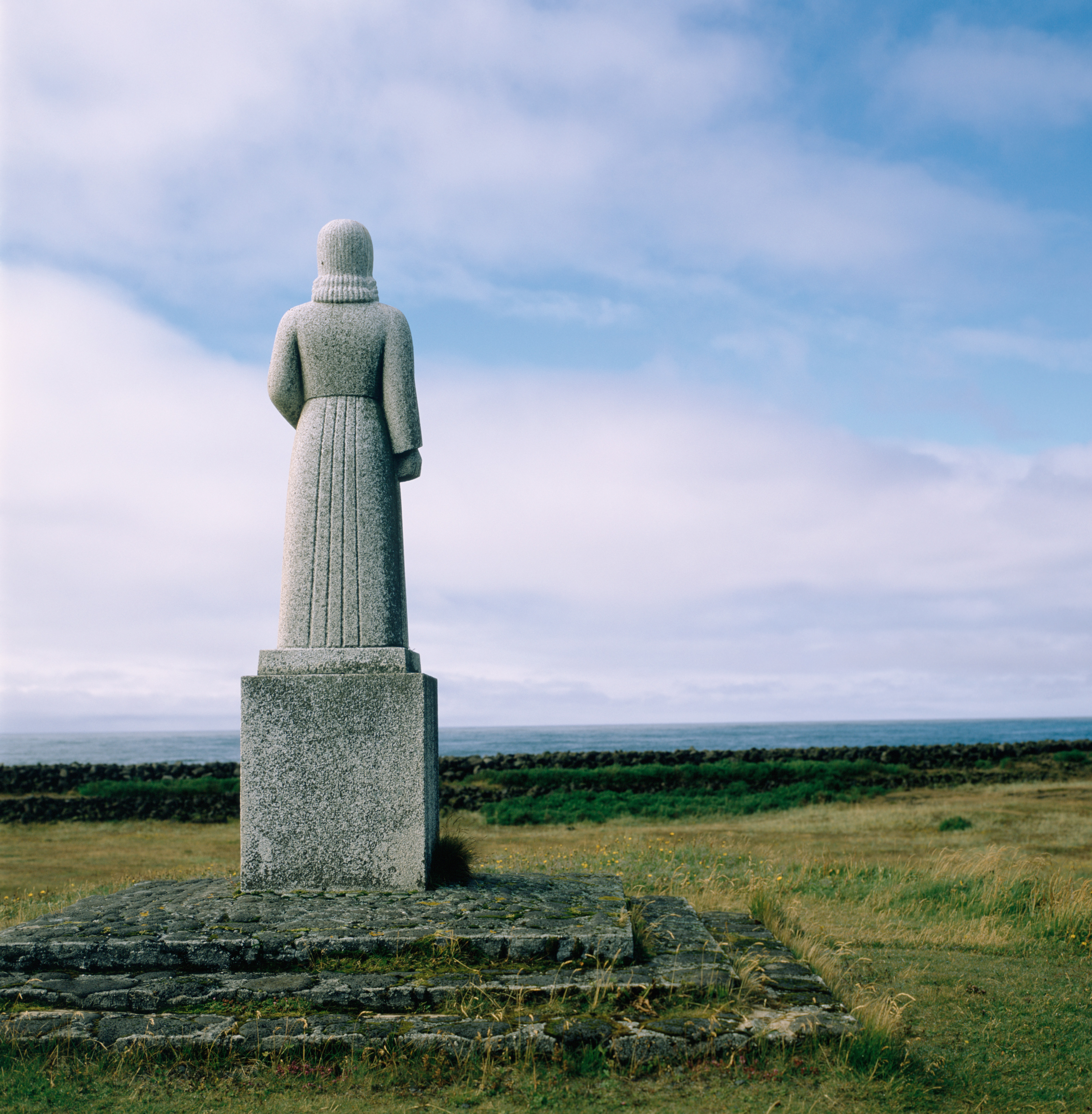 A granite statue of a woman from behind which stands on a small hill. The sky is deep blue with wisps of clouds. Green fields surround the statue.