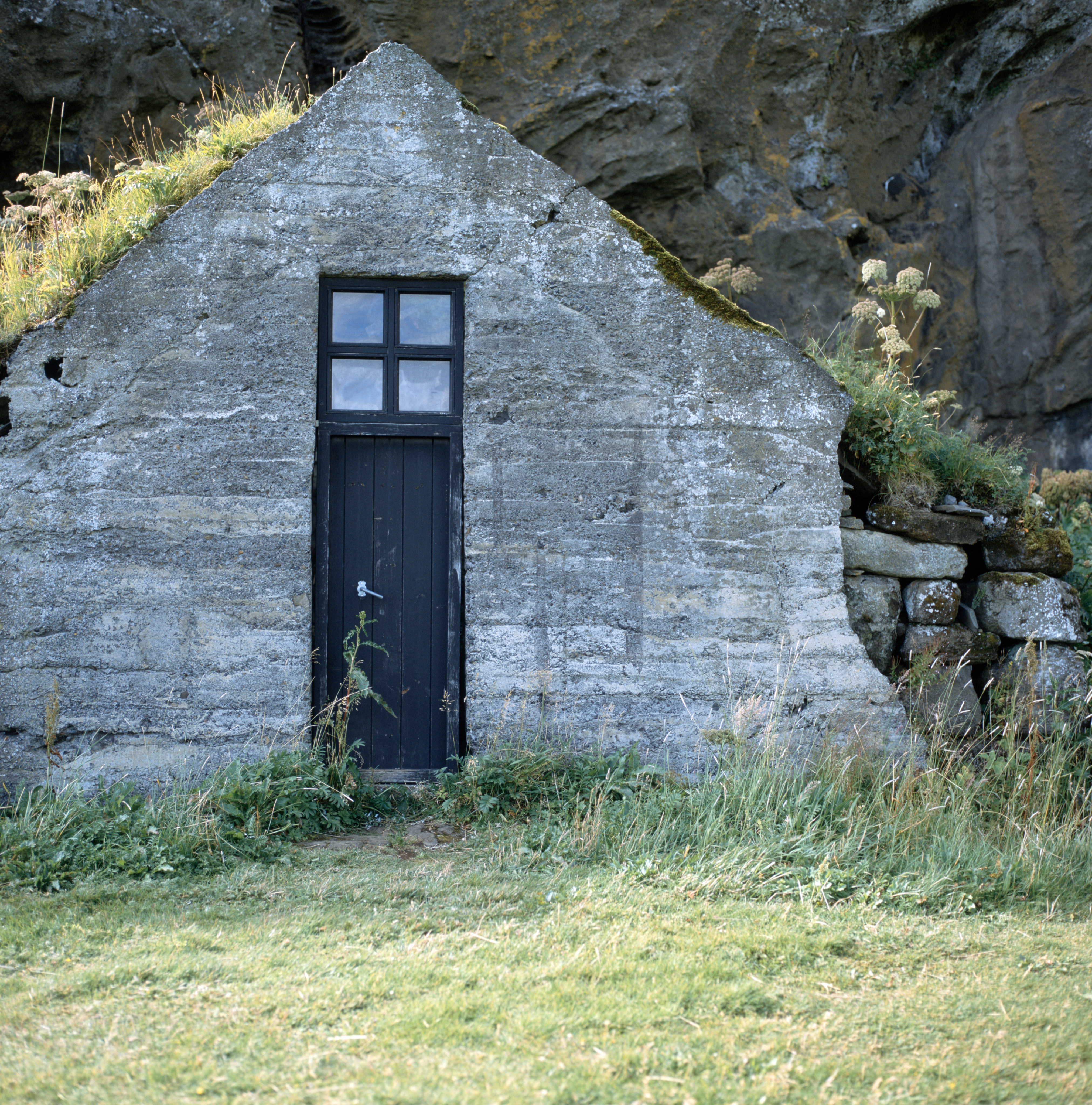 The stone front of a house built into a cliff. There is a black door with a four-paned window directly above the door. The roof is earthen and has grass and other vegetation growing on it.