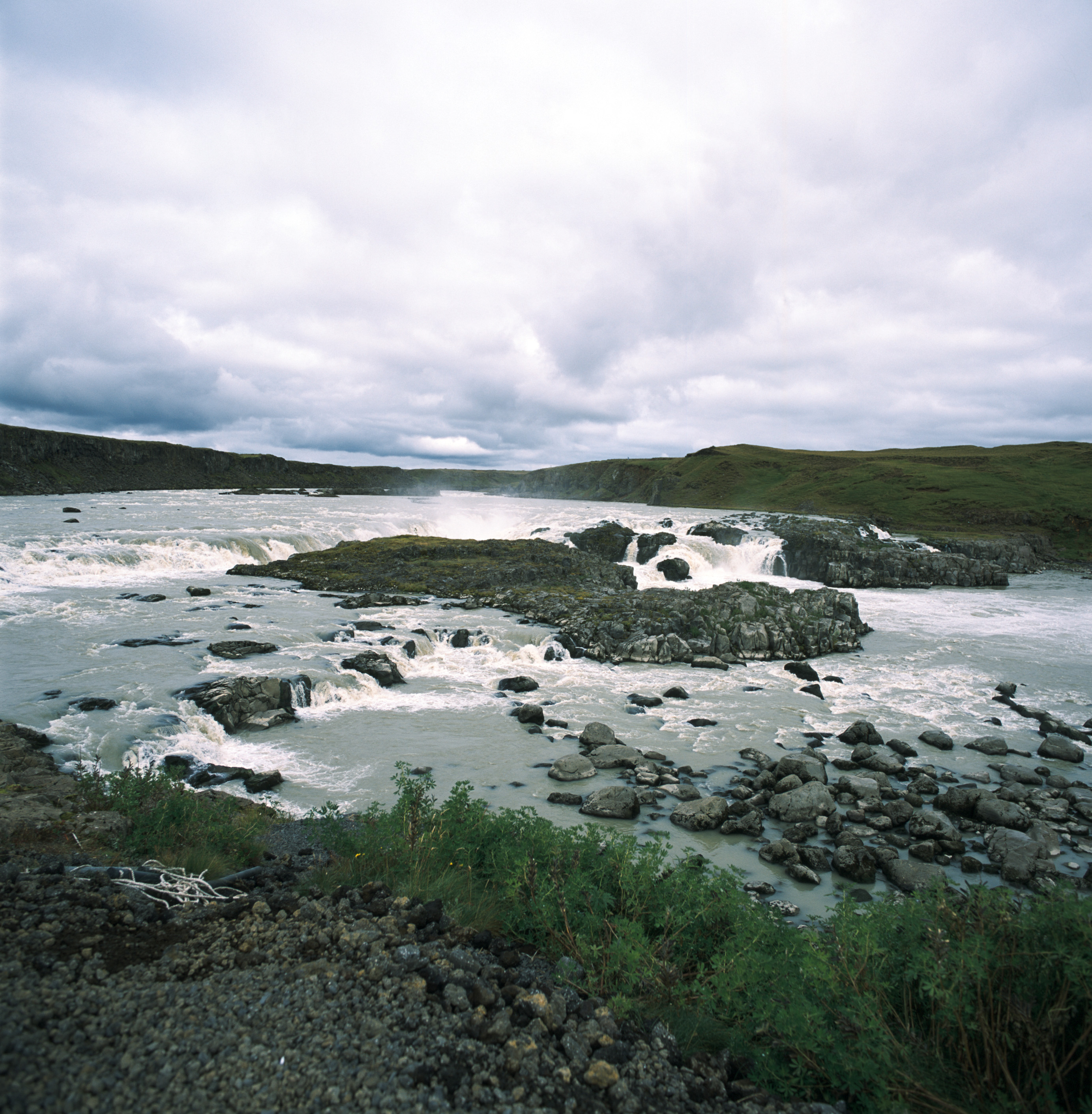 A small, wide waterfall flows over rocks. There are green hills and cliffs in the background and black rocks studded throughout the stream of water. The water is crisply frozen in time.