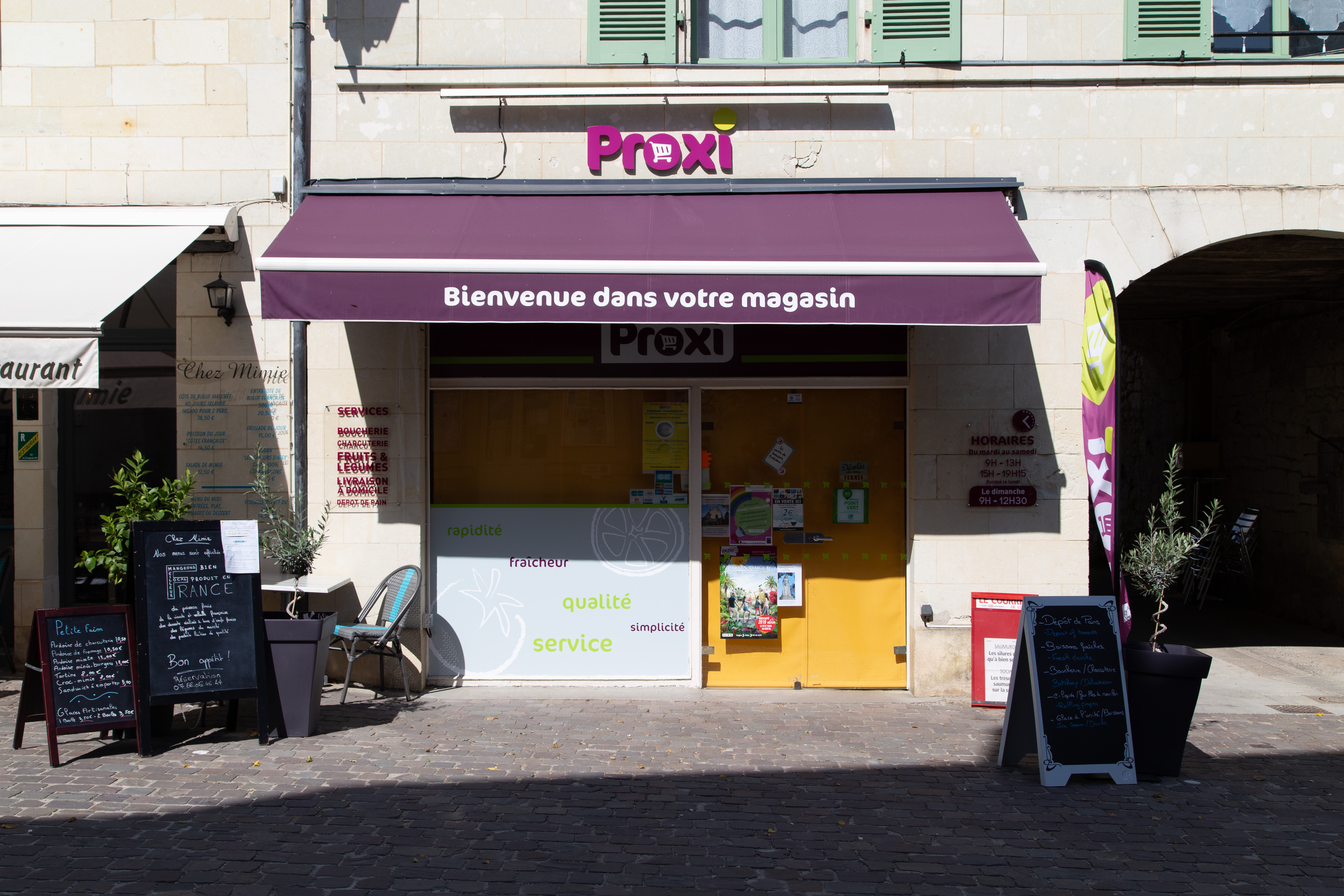 The façade of a small Proxi store, closed for the day. There is a large purple awning that reads "Bienvenue dans votre magasin" below that a window and glass door with a large yellow/orange screen pulled down. Outside, a small table and chairs with menu boards for the restaurant next door on the left.