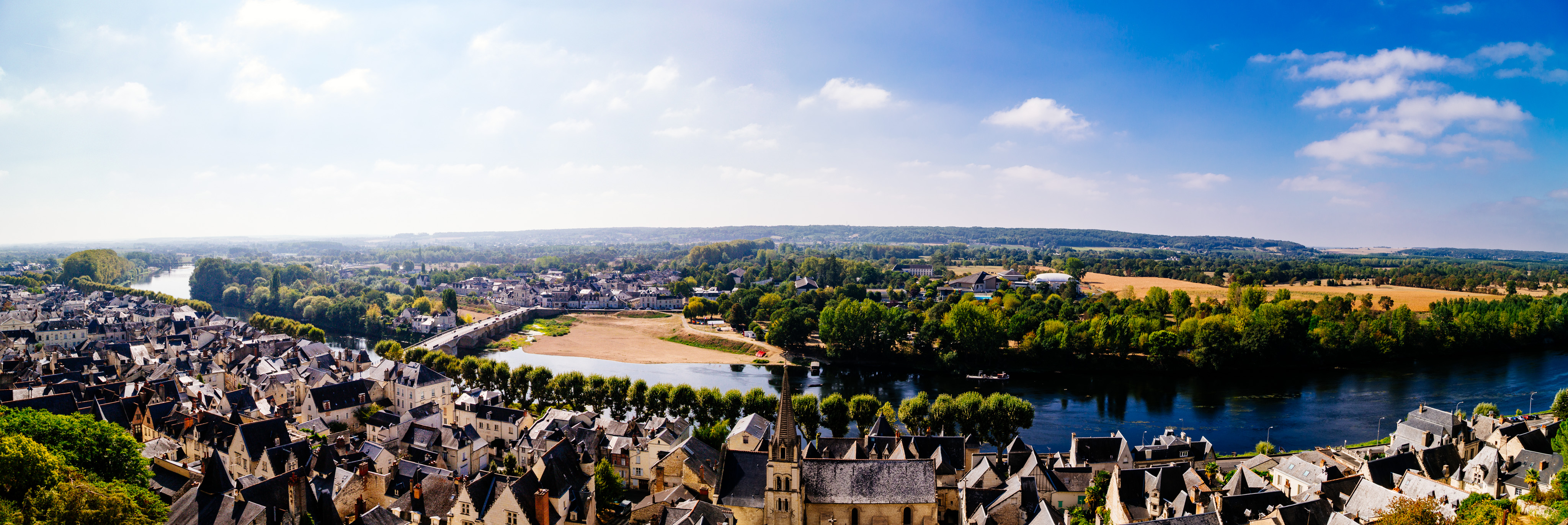 A panoramic view of the Loire river taken from Chinon. The river stretches through the middle of the frame. Trees line the far bank, and a traditional french town stretches down the near bank. In the center a church spire pokes up above the roofs of the 2-3 floor buildings that cover the foreground.