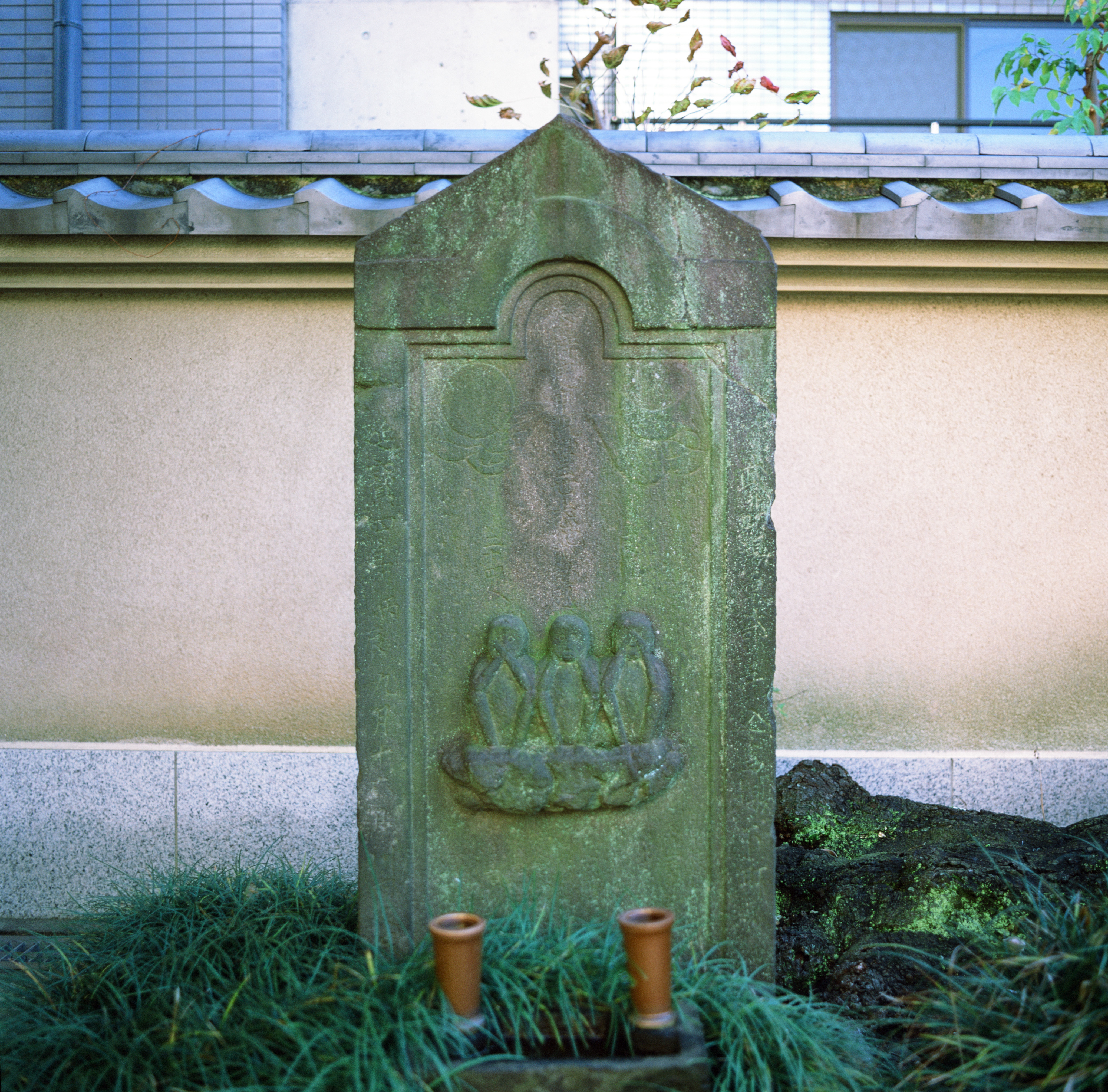 A stone monument, green with moss growing on it. In the center of the stone is a relief of the three wise monkeys (speak no evil, hear no evil, see no evil). Behind the stone is a beige wall with gray tiles on top. In front of the stone are two bronze-colored flower holders and plants.