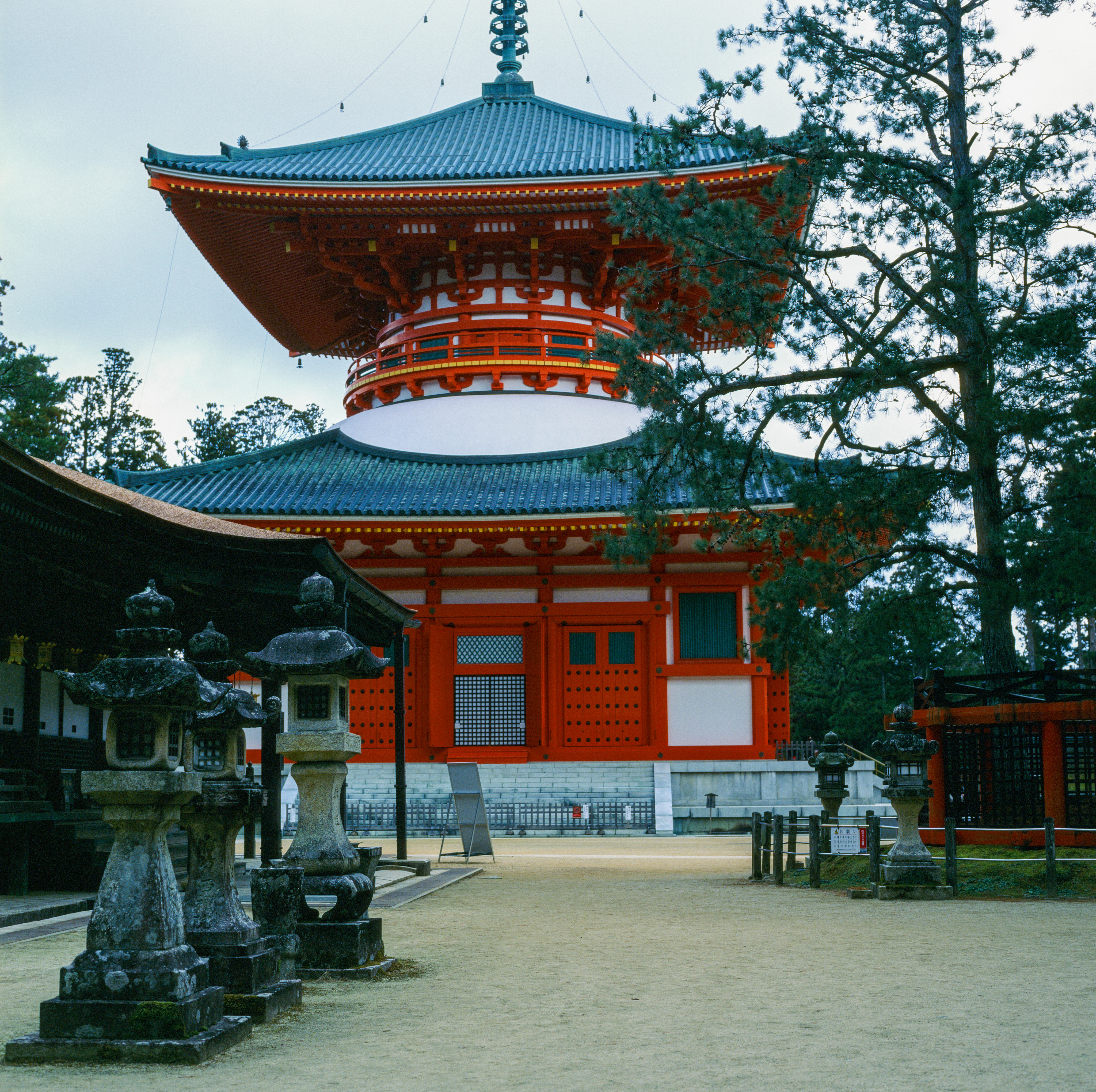 A large red, white, and black pagoda. In front of the pagoda to the left is a line of three stone lanterns, to the right is a large pine tree. The pagoda has two levels, with a spire at the top.