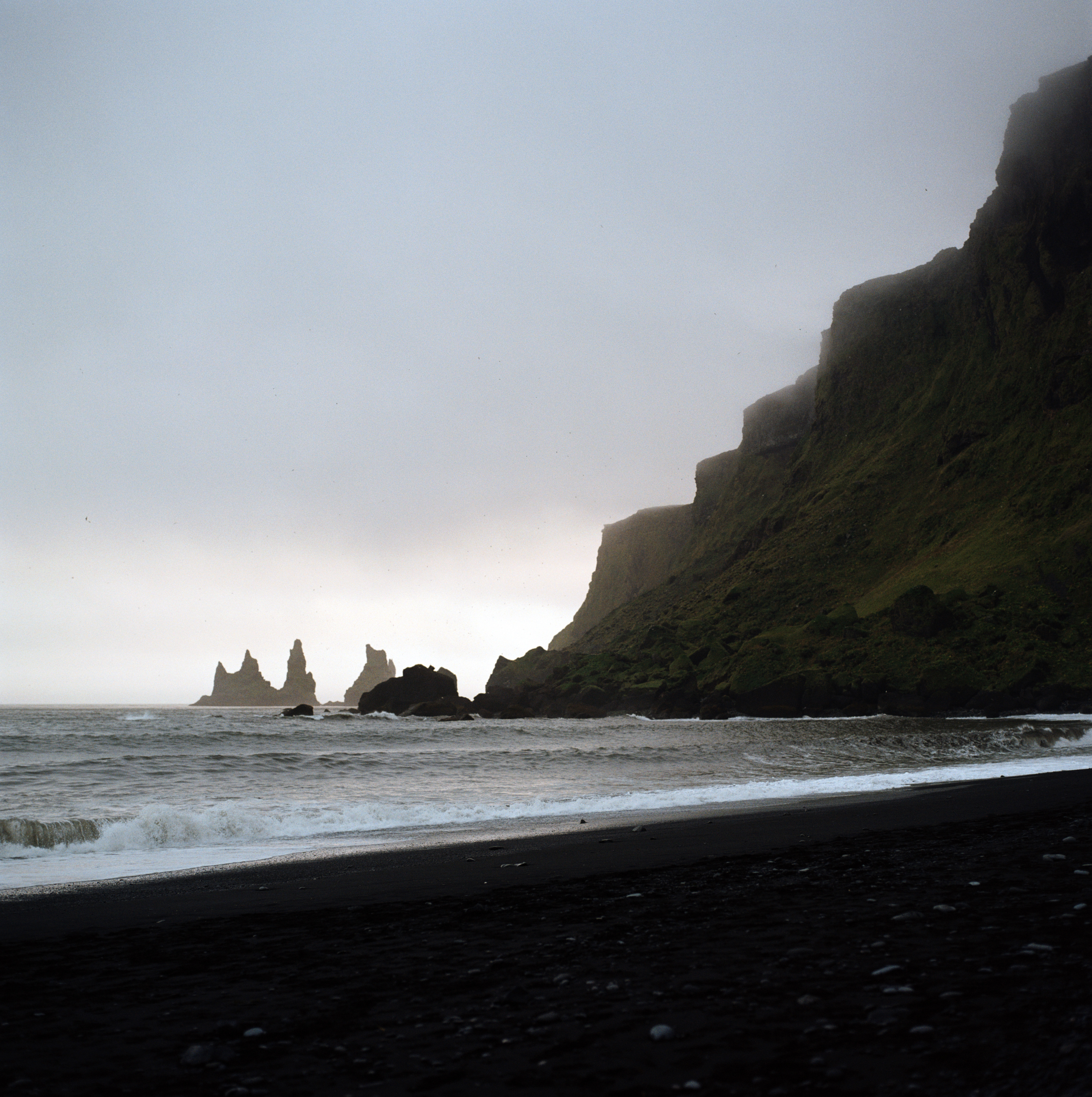 A diagonal line of cliffs and islands goes from the top right of the frame to the bottom left. Starting with cliffs on the land and leading down to jagged small islands. Water is rolling in towards a black sand beach in the foreground.