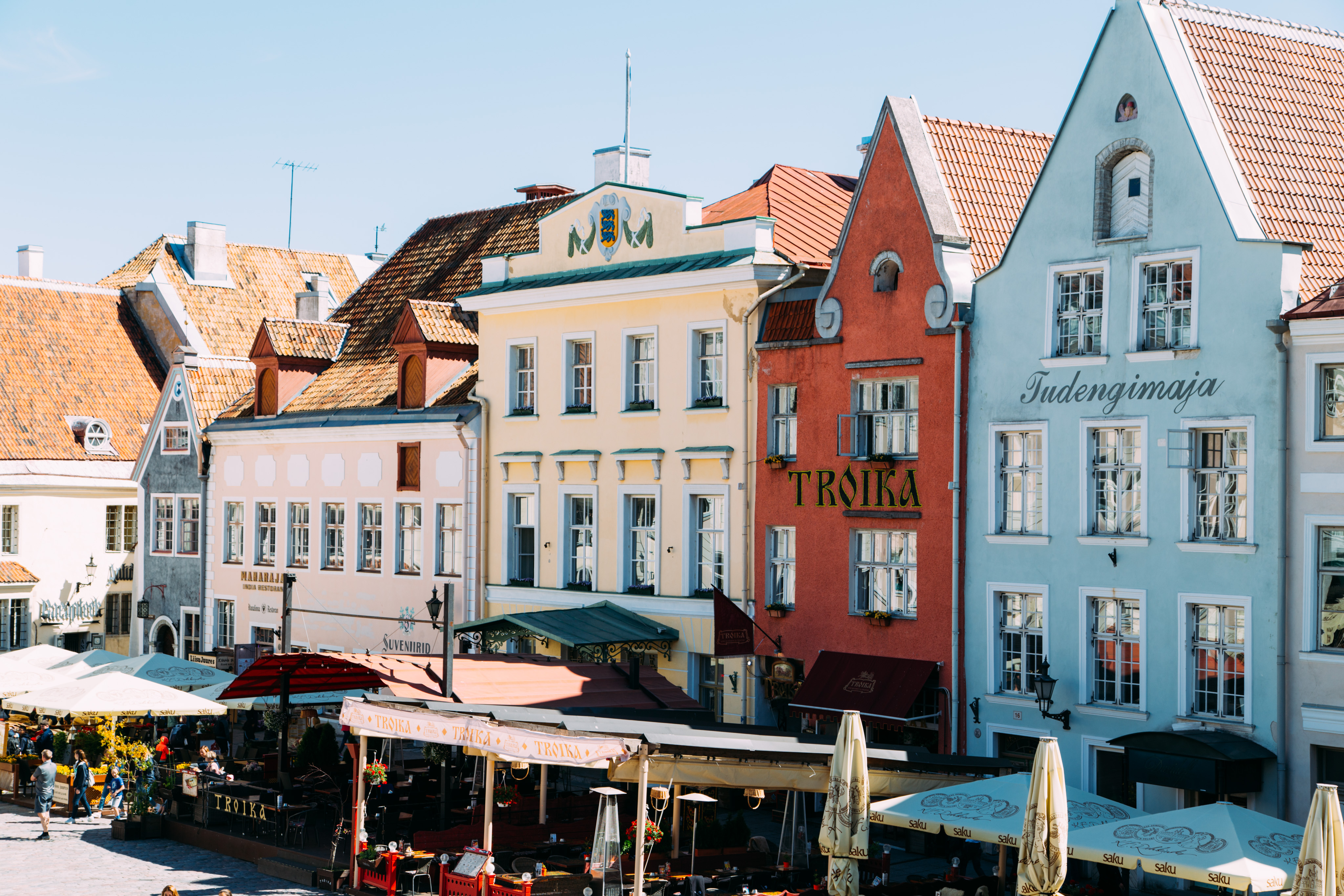 A string of buildings in the Old Town Square (Raekoja Plats) in Tallinn, Estonia. The buildings are blue, pink, yellow, burnt orange, and blue from left to right. The last two buildings on the right have the text "Troika" and "Tudengimaja" on them. Awnings cover cafe seating below the buildings.