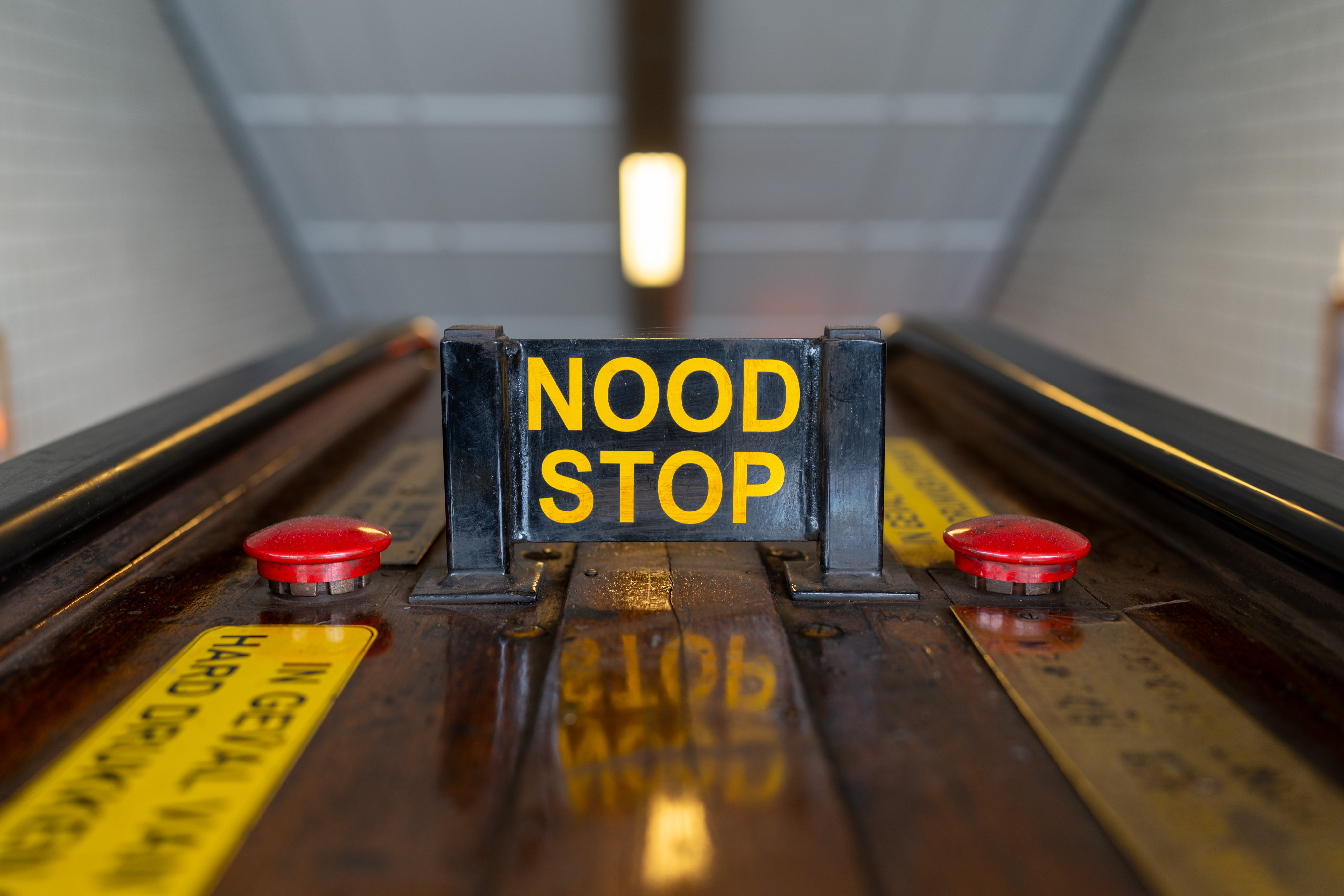 A sign on an escalator that reads "nood stop"  in yellow letters. "Nood stop" means "emergency stop" in Dutch. There are two red buttons on either side of the sign.