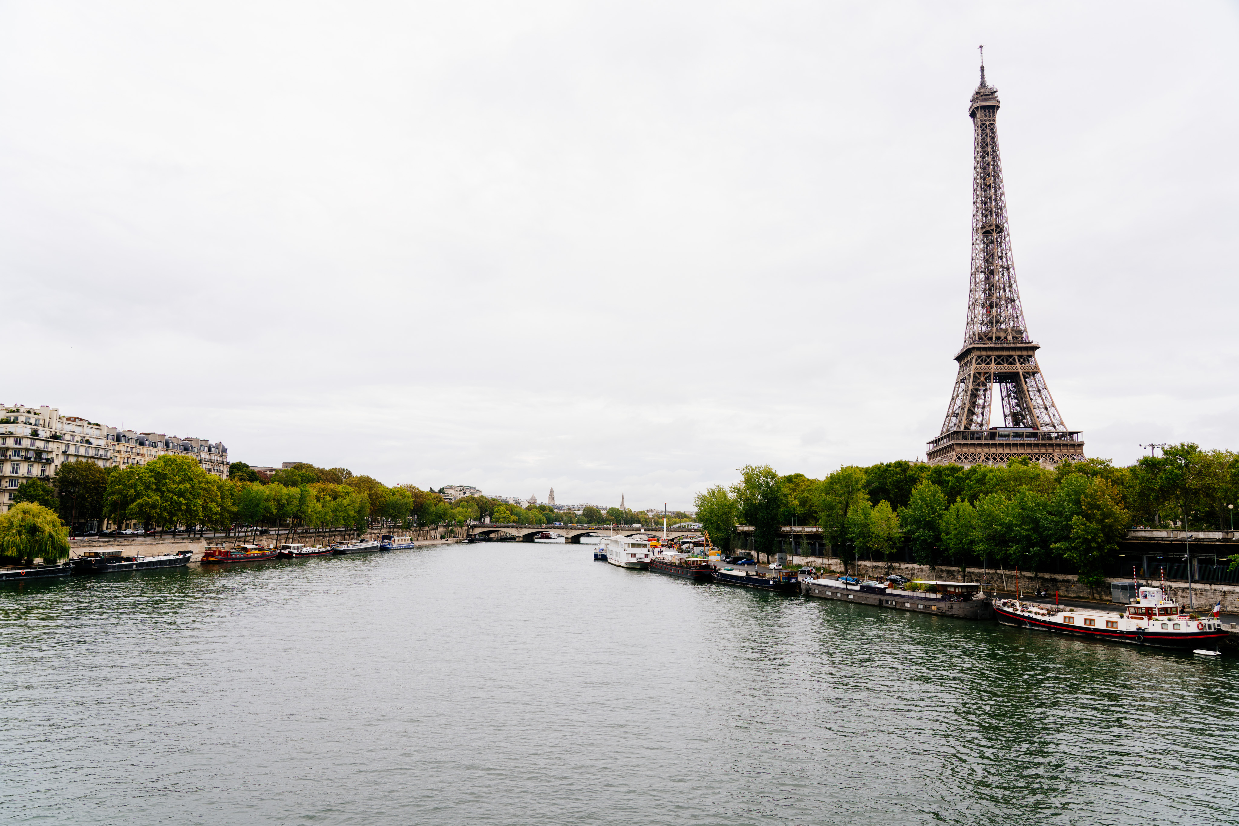 The Eiffel tower stands tall on the right side of the photo. The Seine is in the center of the frame with a number of medium sized boats lining the bank near the Eiffel Tower. A bridge can be seen in the distance.
