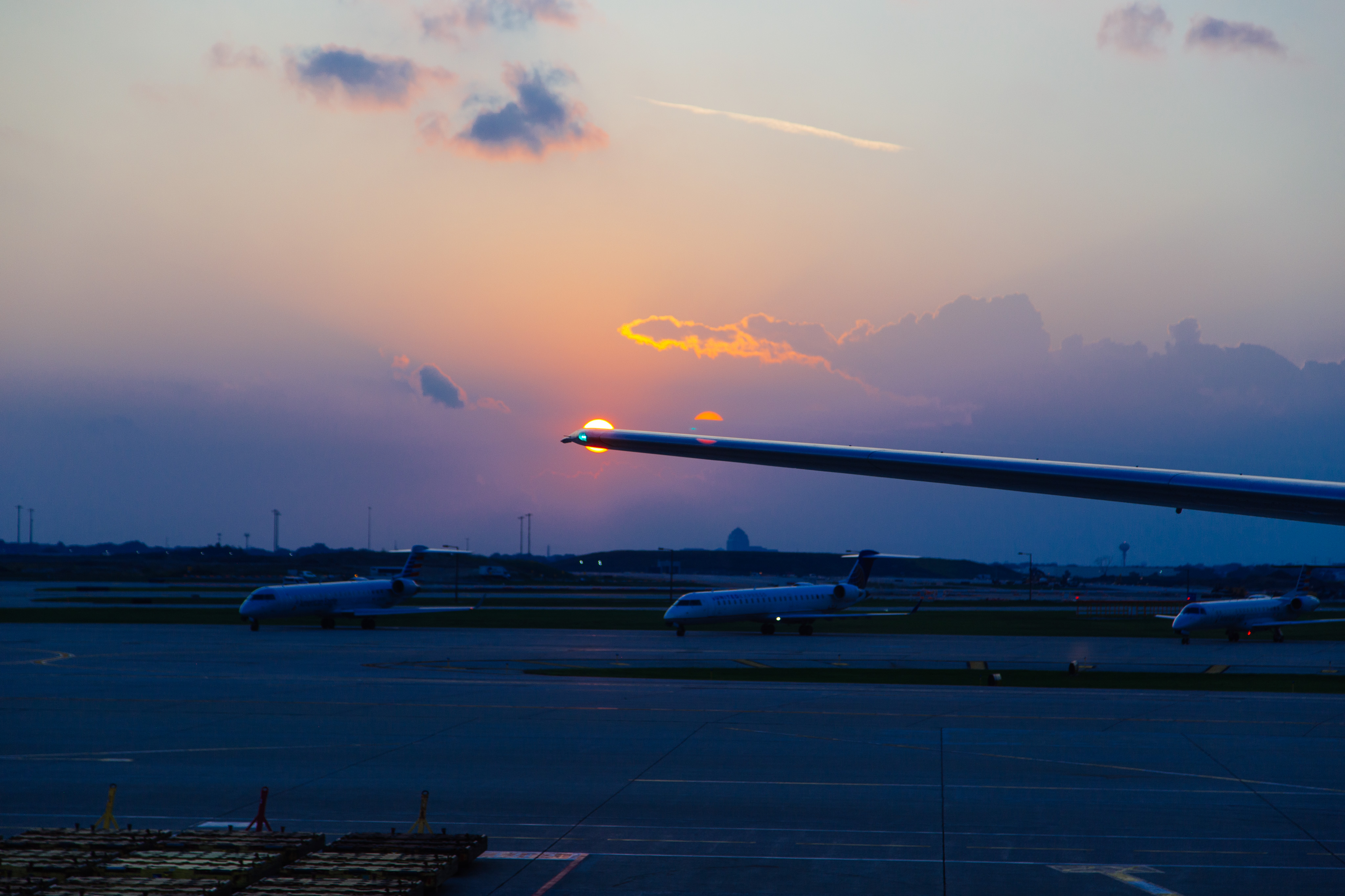 A 777 wing is partially obscuring the setting sun. A ghost of the sun and the wing light can be seen next to it. Three smaller regional jets are parked in the background.