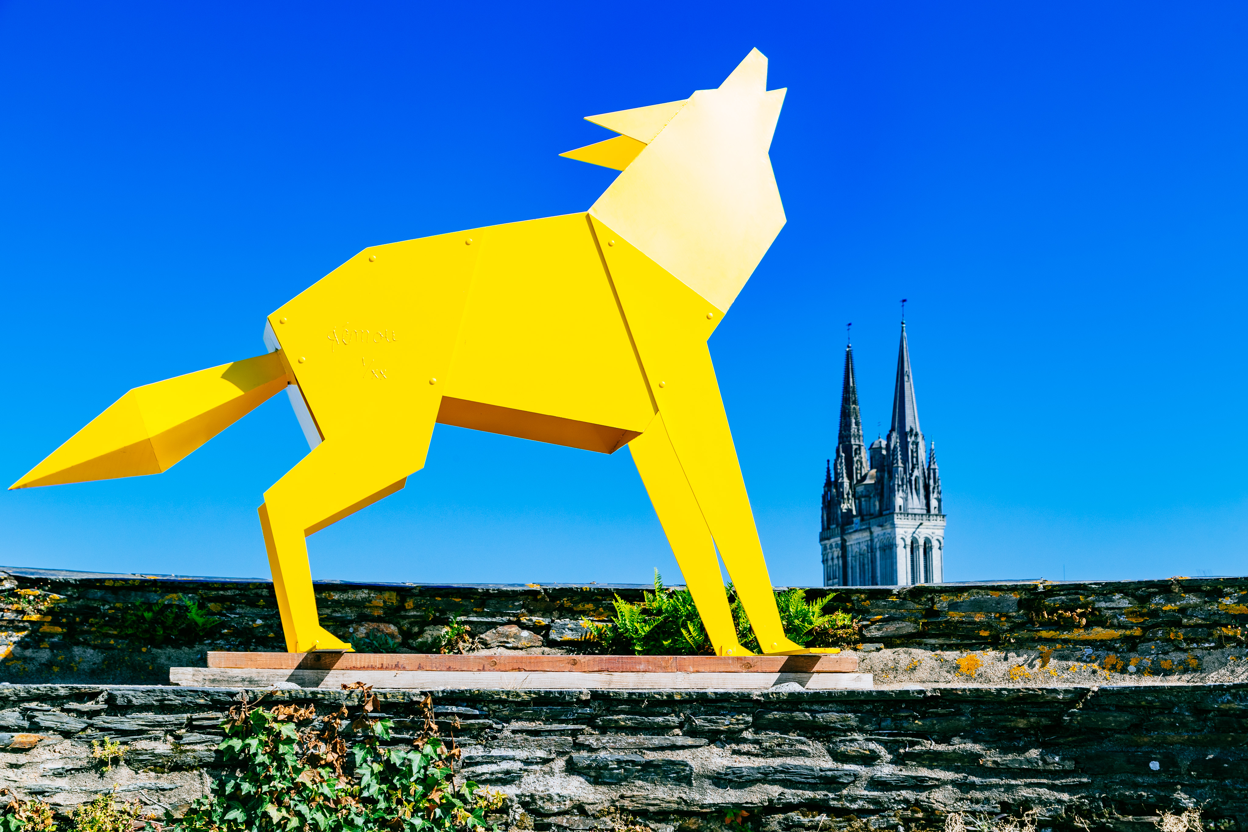 A large yellow metal sculpture of a wolf howling looms large. The statue is reminiscent of origami which sharp angles. To the right of the wolf are two towers of a cathedral