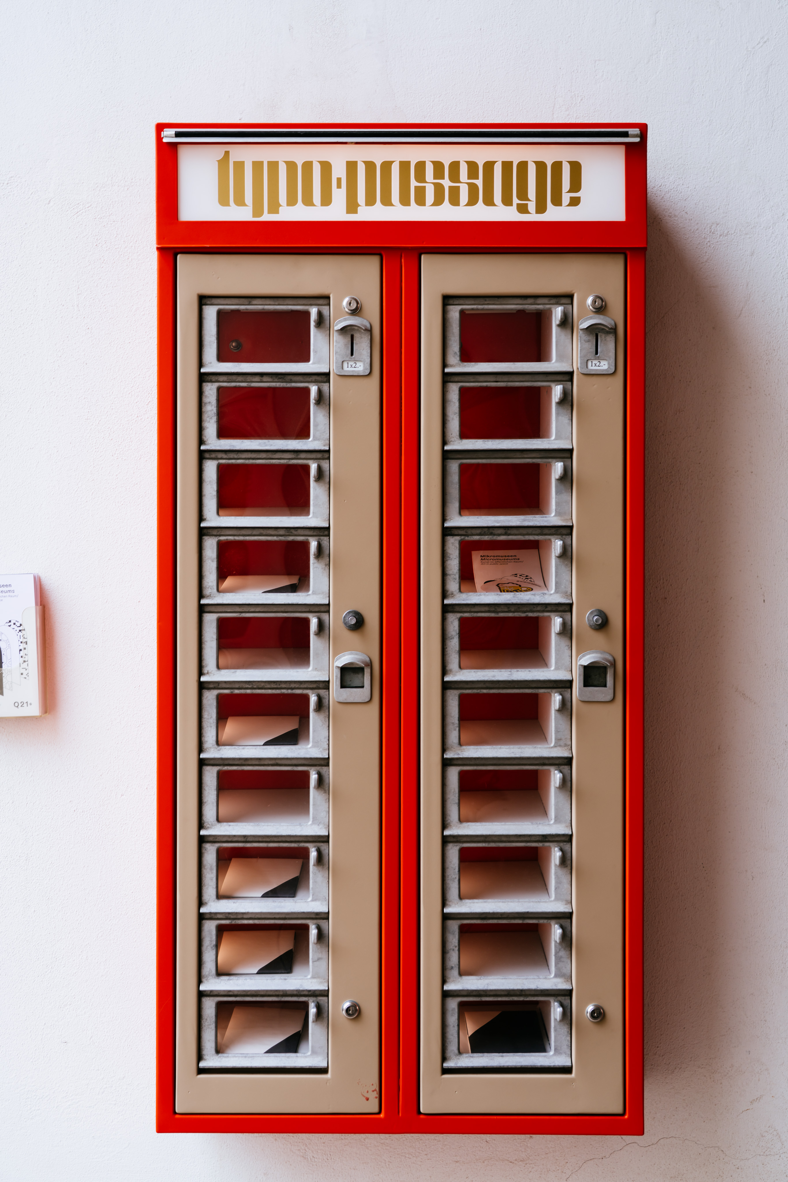 An old fashioned vending machine. The machine is red with cream accents. It's label on top says "typo-passage". There are two columns of ten doors with windows. In a handful of the windows are small pamphlets.