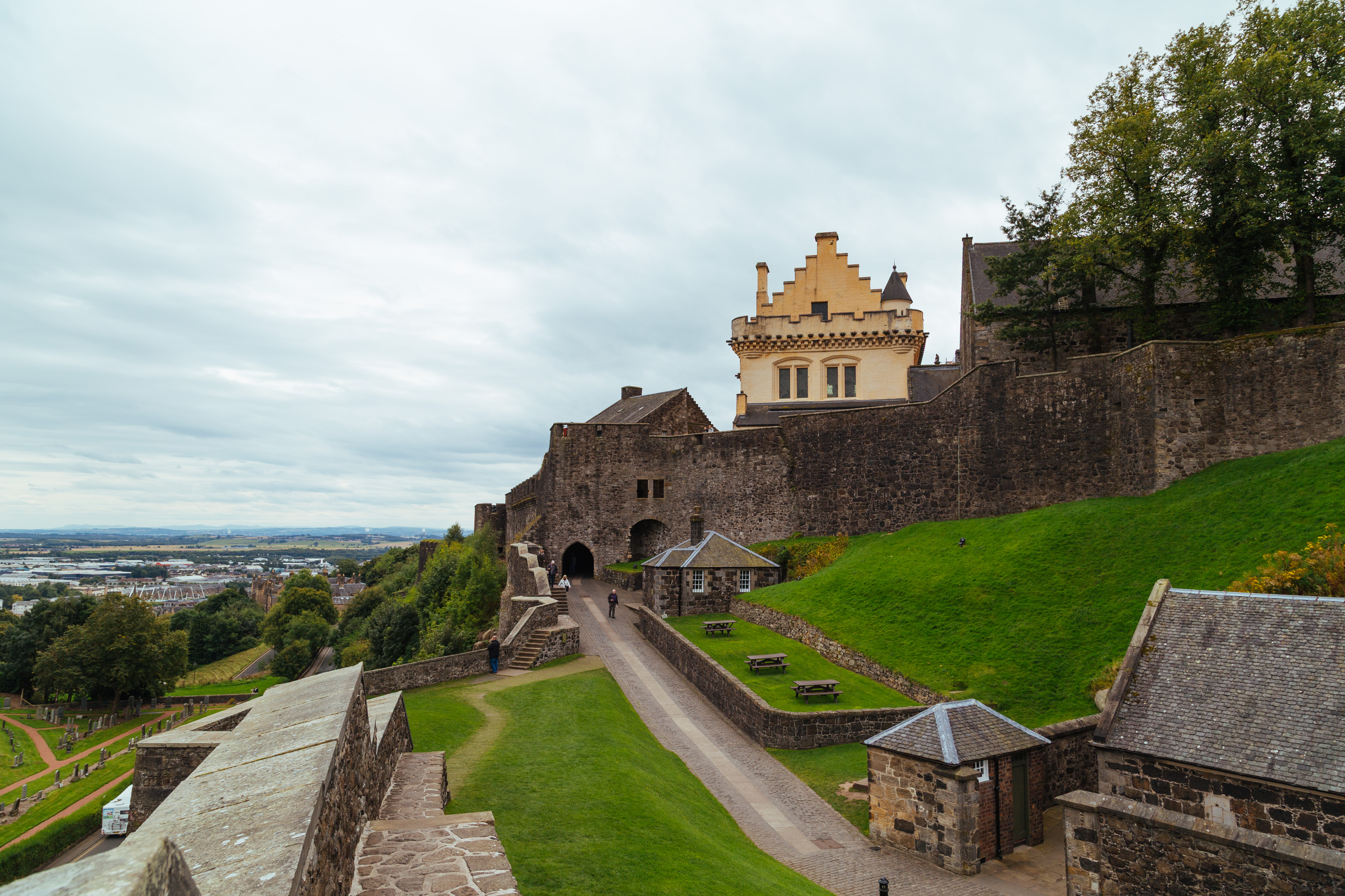 A view inside of the Stirling castle grounds. A terraced area rises up from the left of the photo to the right. From the left, there is a wall, then a small road, a few small buildings, a terraced park and a sloping hill up to another (inner) wall. The main road leads to a small arched portal. Just above and to the right of this portal rises a central building at Stirling castle. This building is colored a light yellow, indicative of how it would have looked when it was first built. In the background in the left of the photo is the sprawling town around Stirling.