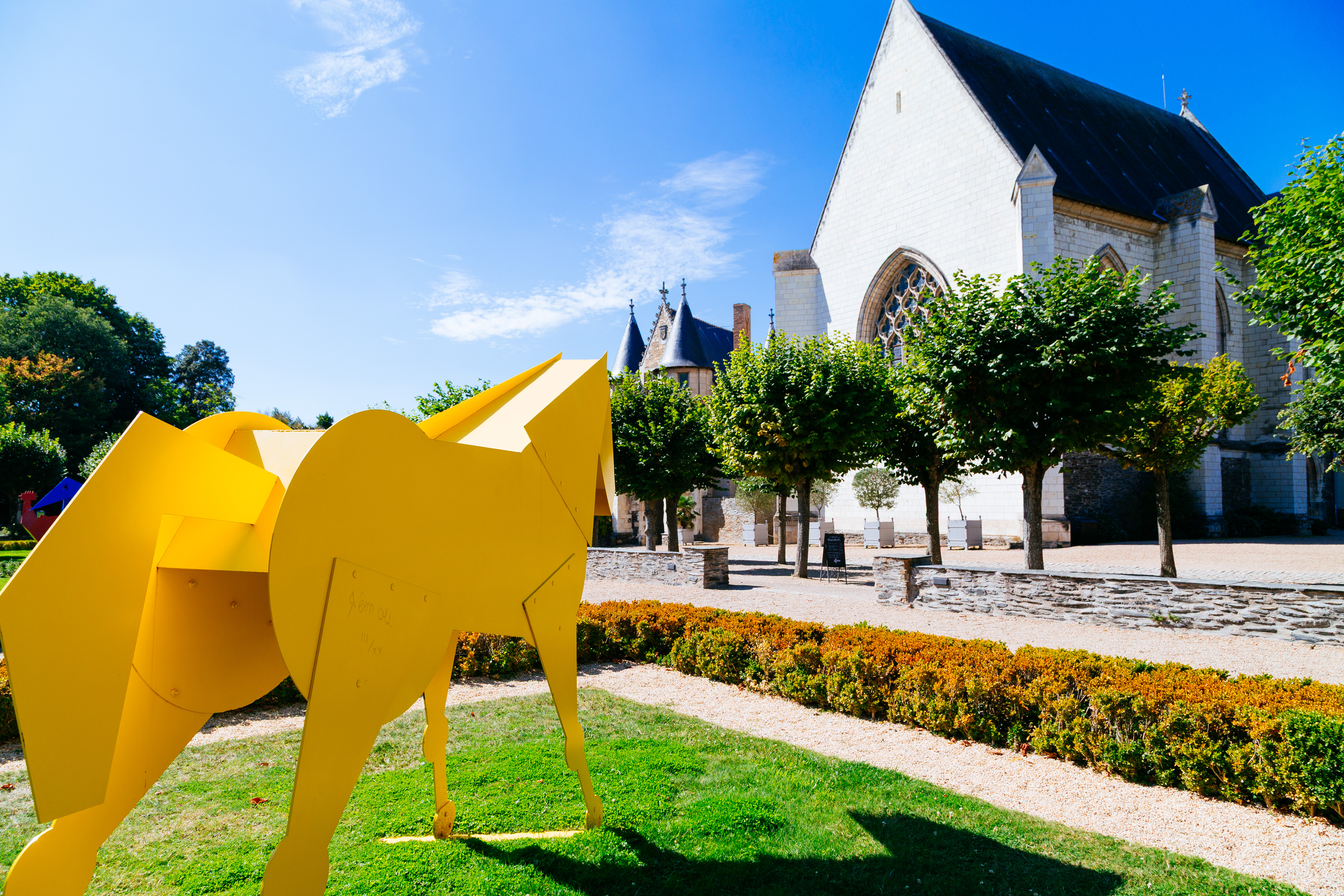A large yellow metal sculpture of a horse stands facing a chapel building. Between them is a row of trees and a manicured garden.