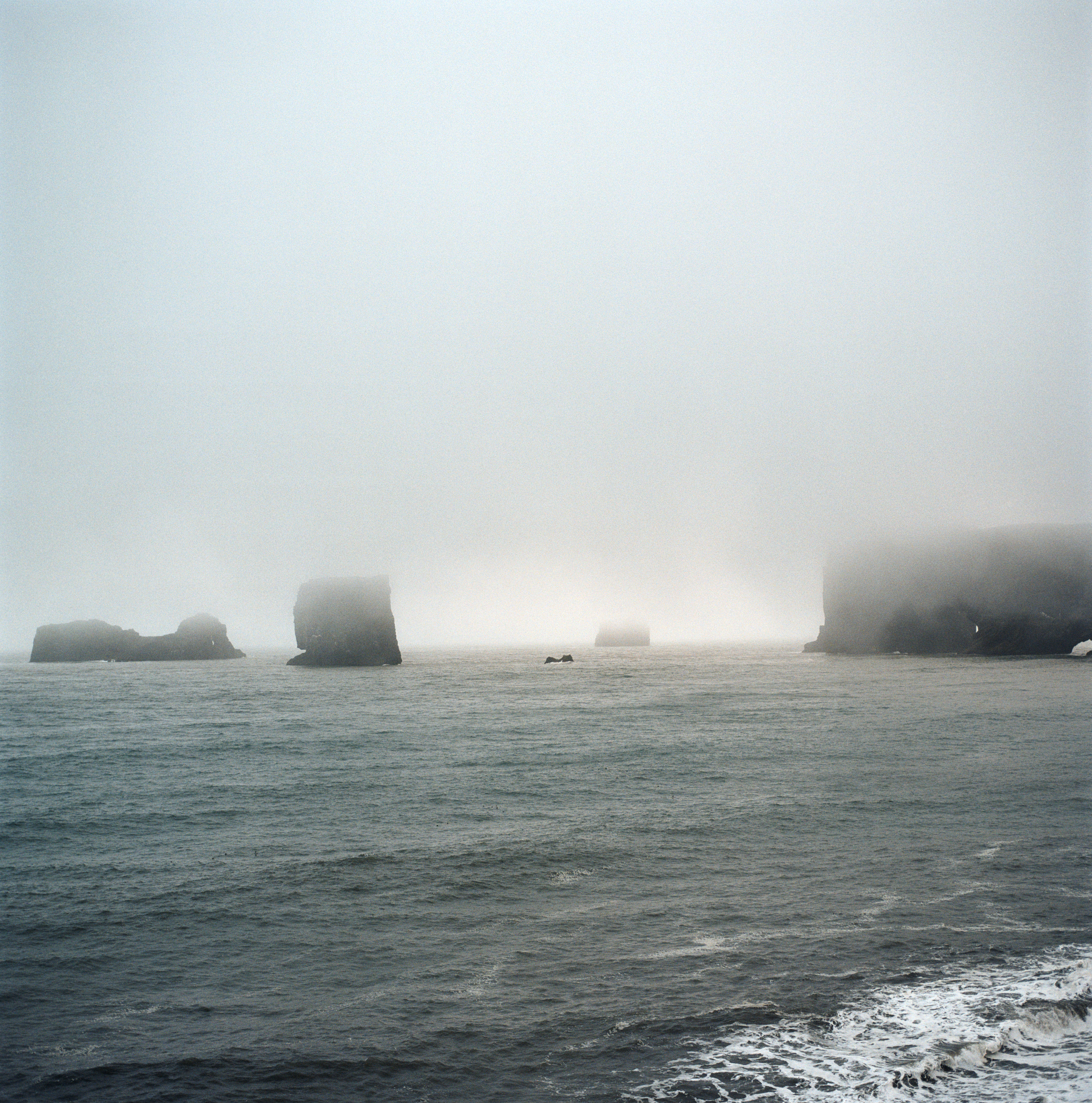 A set of small, black islands in the see are engulfed in the top half of the frame by fog and clouds. there are gentle waves crashing in the near-foreground