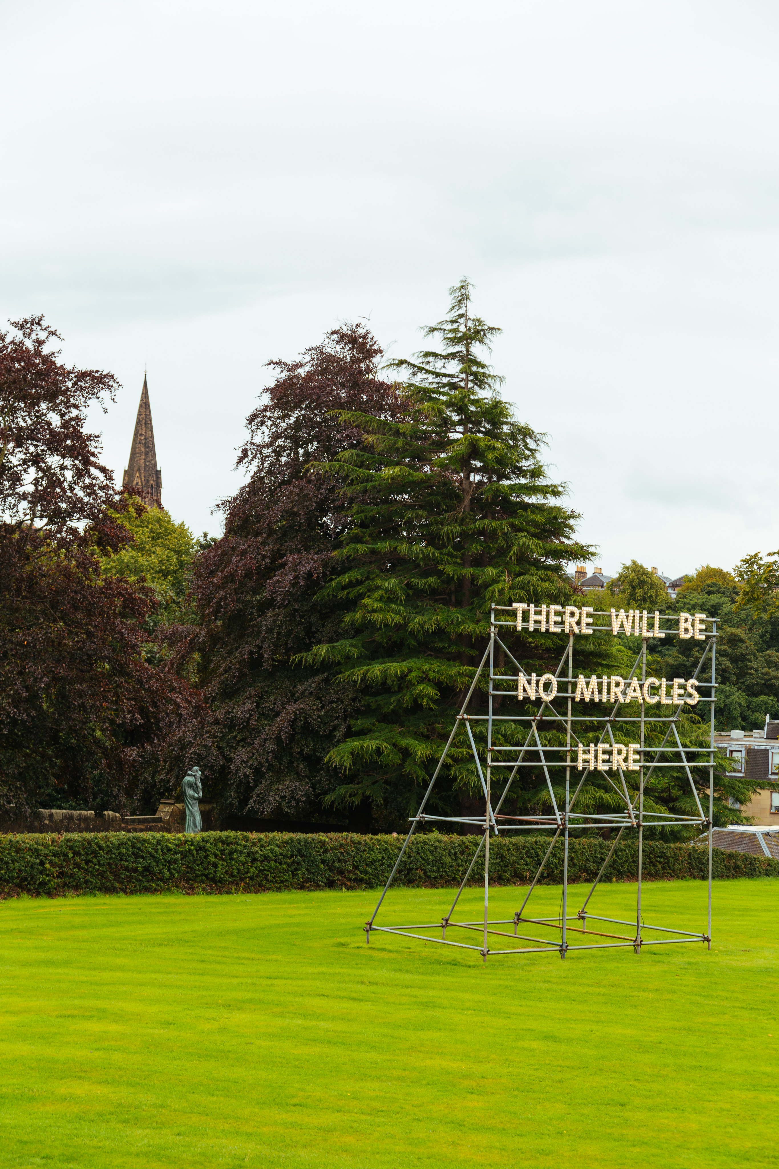 A view onto the yard of the Scottish National Gallery of Modern Art. There is a well-manicured lawn with a large metal framework, one which is hung a light-up sign that reads "There will be no miracles here". Behind the sign is a long, square hedge, a small statue and some trees. Just beyond the trees a steeple of a church building pokes up above the trees