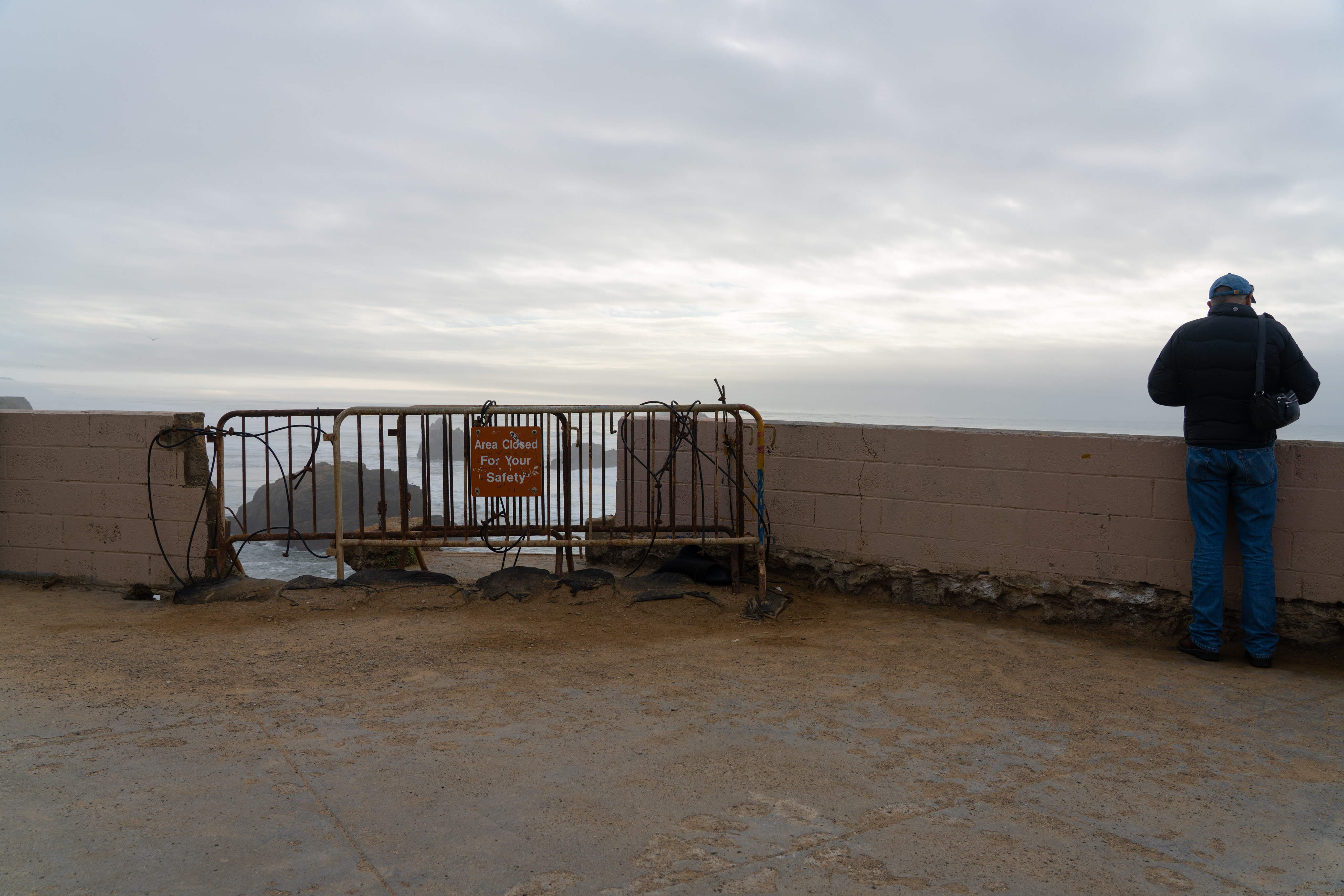 A brick wall with a break in the middle, temporary metal barricades blocking the entrance. On the barricades is a sign reading "Are Closed For Your Safety". A man stands looking out into the distance. In the background the Pacific Ocean is barely visible