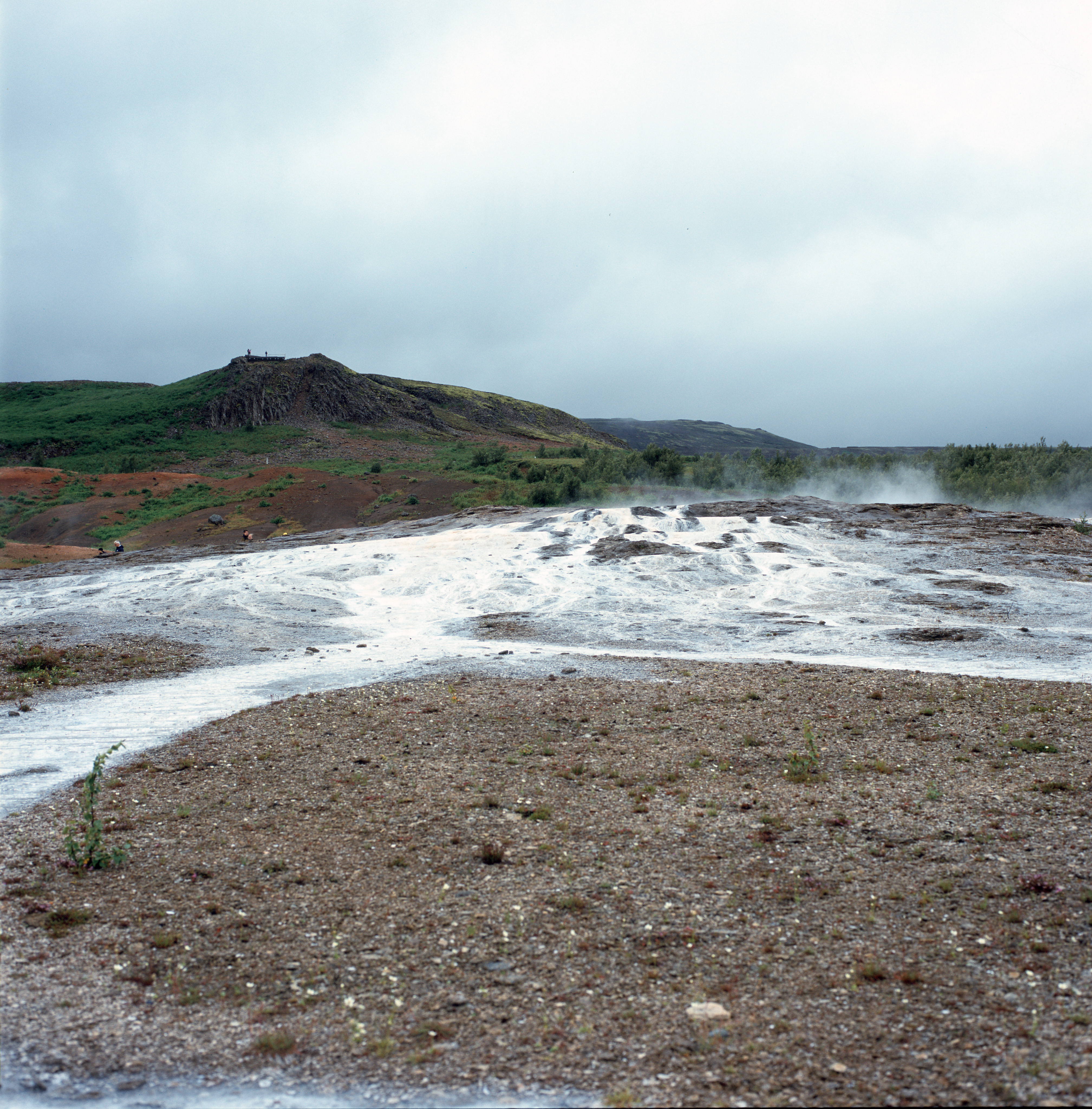 A broad view of thermal fields. In the background a green and black hill stands, in the mid-ground is a wrap of white minerals leading to a rocky and grey foreground. Steam is gently rising in the back to the right.