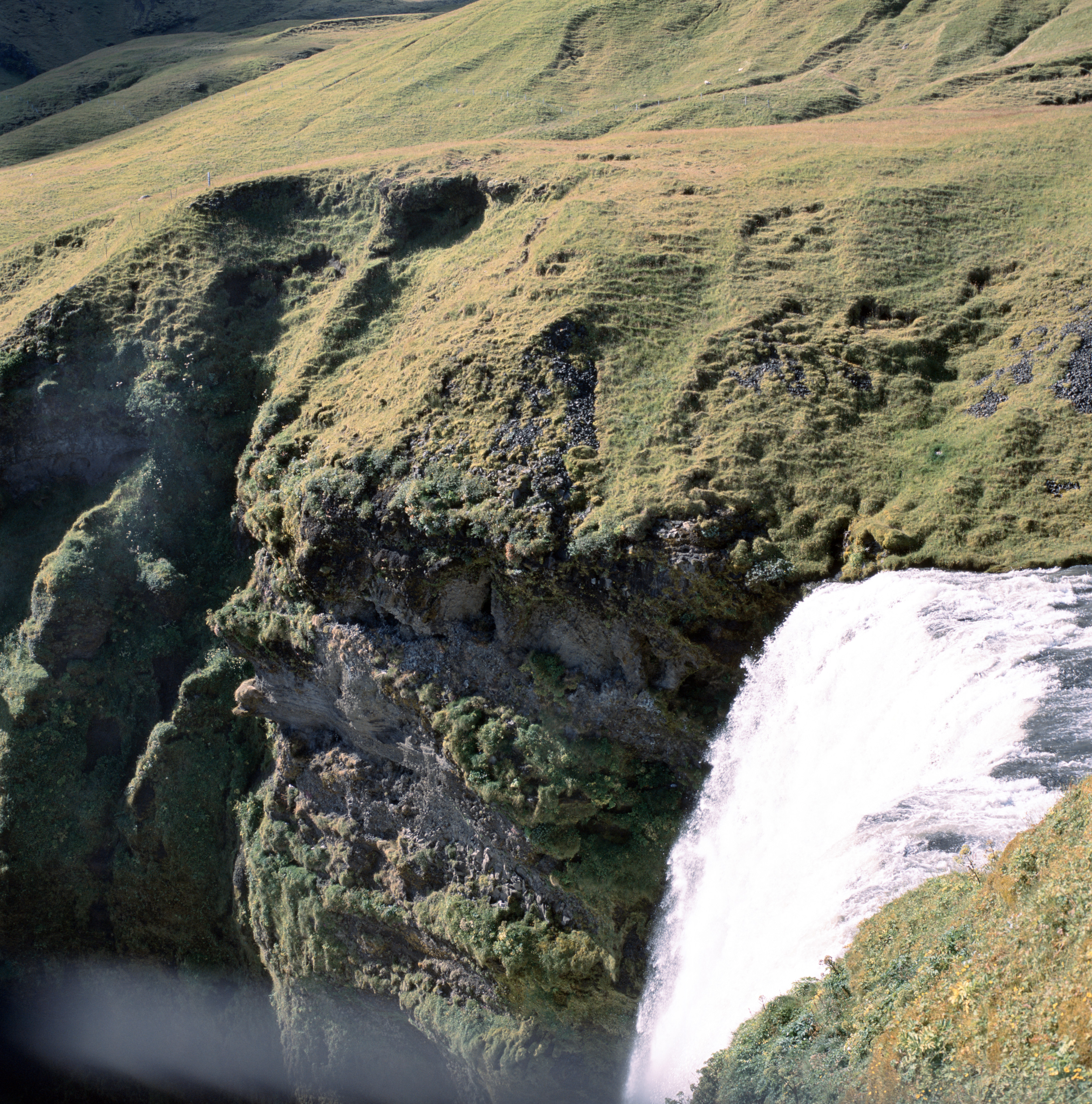 A waterfall flows down in the lower half of the frame. Above and around the waterfall are cliffs covered with green grass and moss.