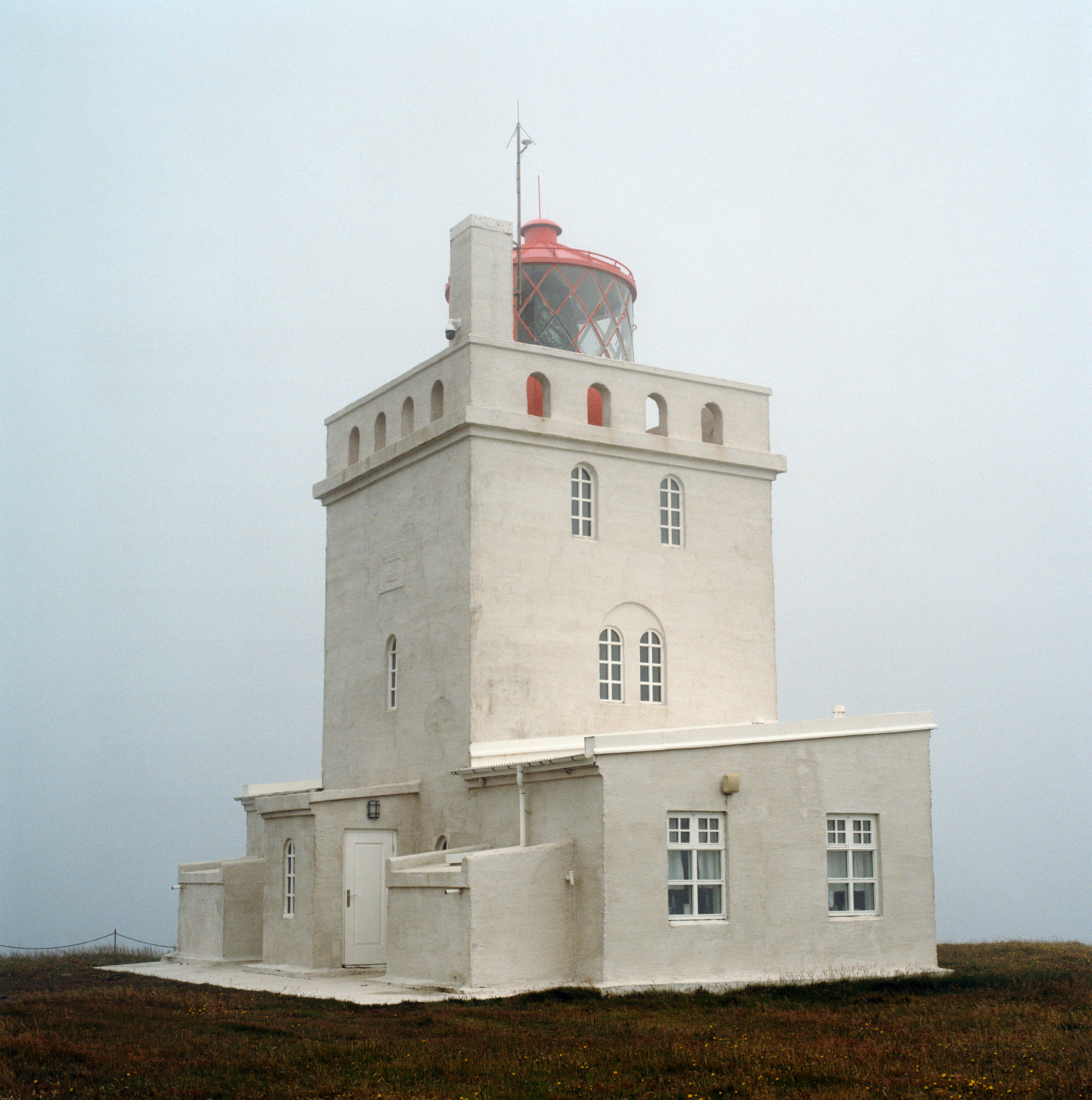 A lighthouse in fog. The lighthouse is white, with a central square tower and some lower extensions on either side. The light is housed in a red metal and glass enclosure.