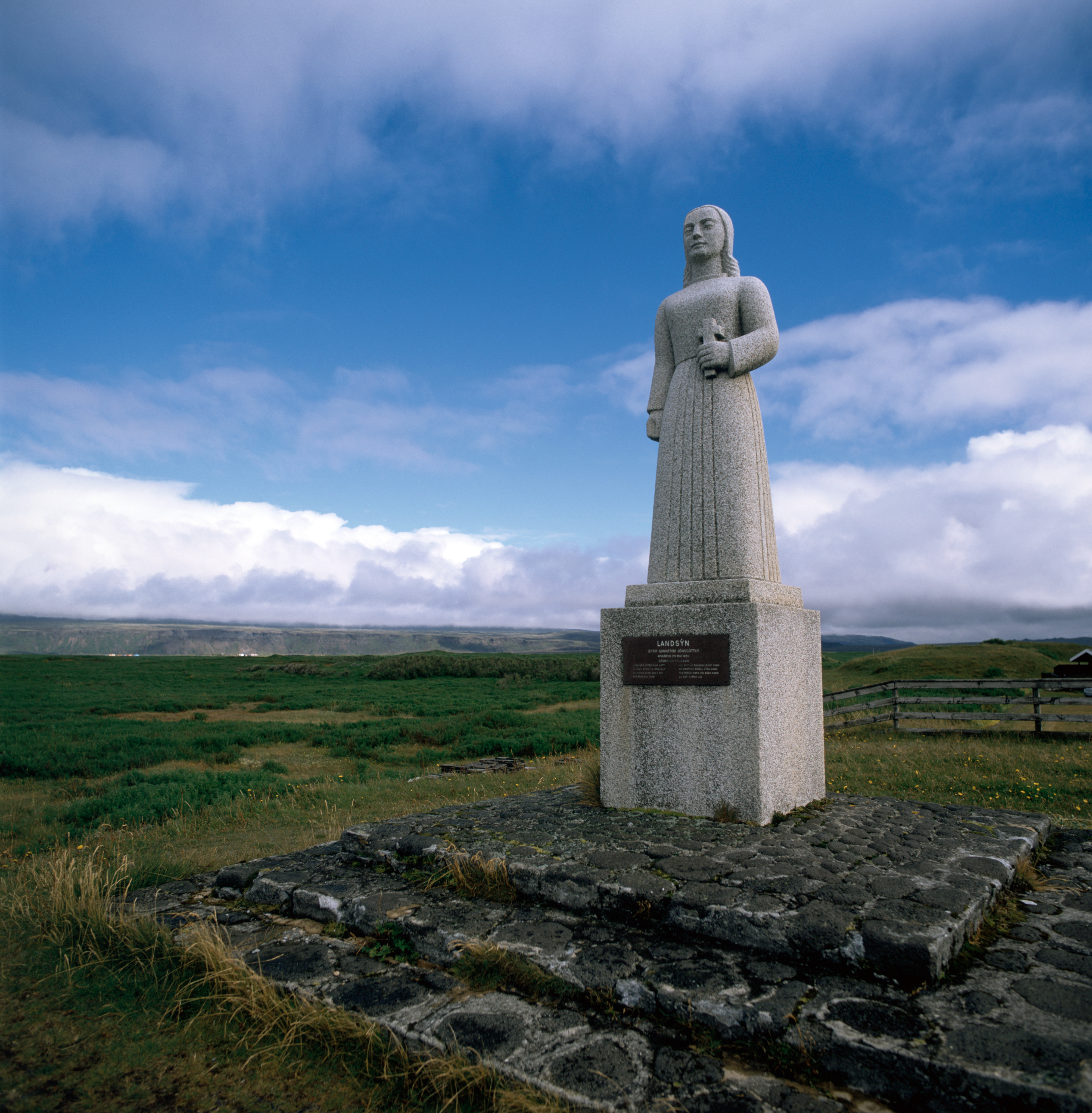 A granite statue of a woman holding a cross stands on a small hill. The sky is deep blue with wisps of clouds. Green fields surround the statue.