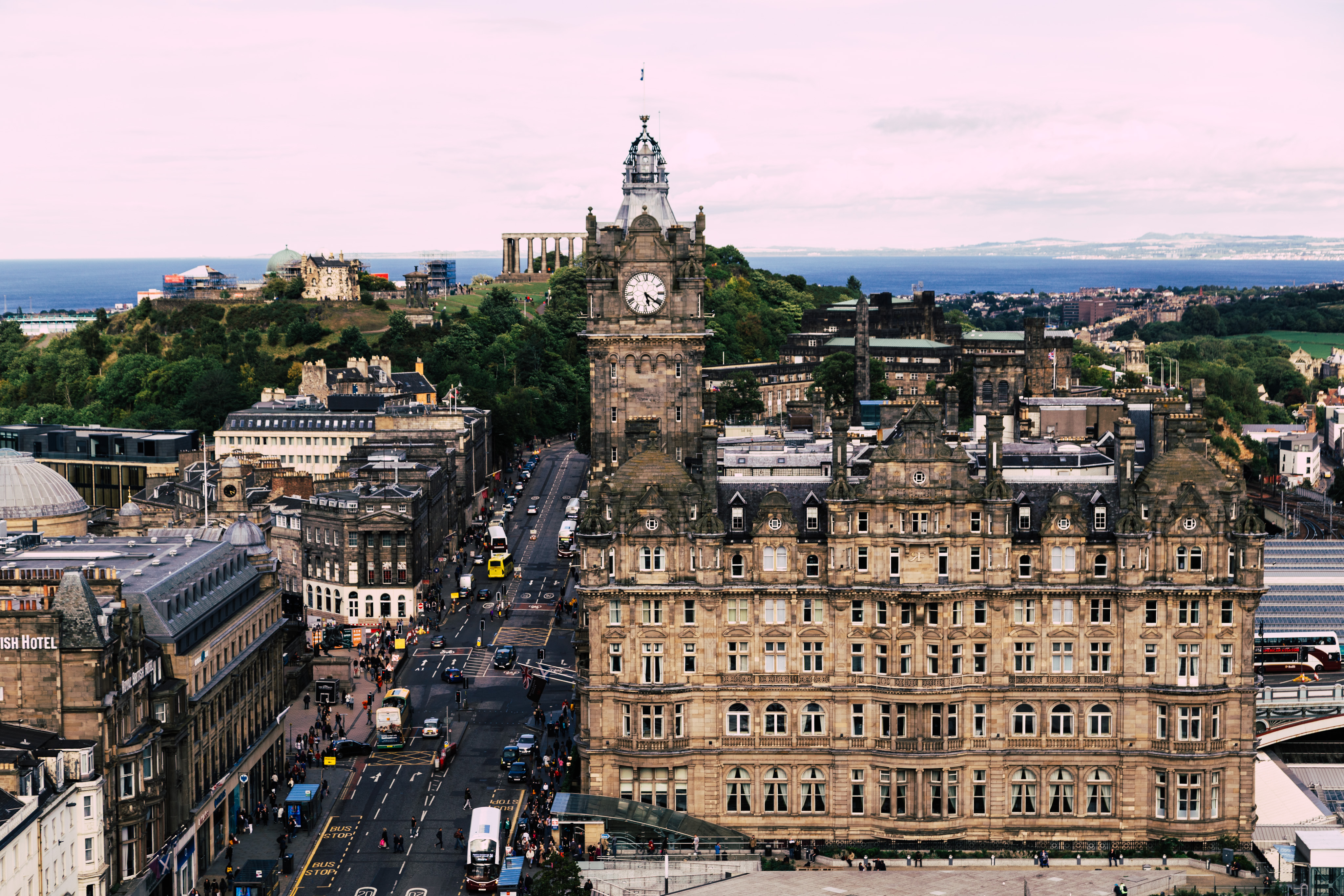 A view from the Scott Monument in Edinburgh. The bulk of the frame is the large Waverley mall, a busy street runs alongside it continuing on into the distance. In the background, to the left fo the building is Calton hill which contains a number of monuments and other buildings. Distant in the background is the Firth of Forth stretching the width of the frame, distant in the have.