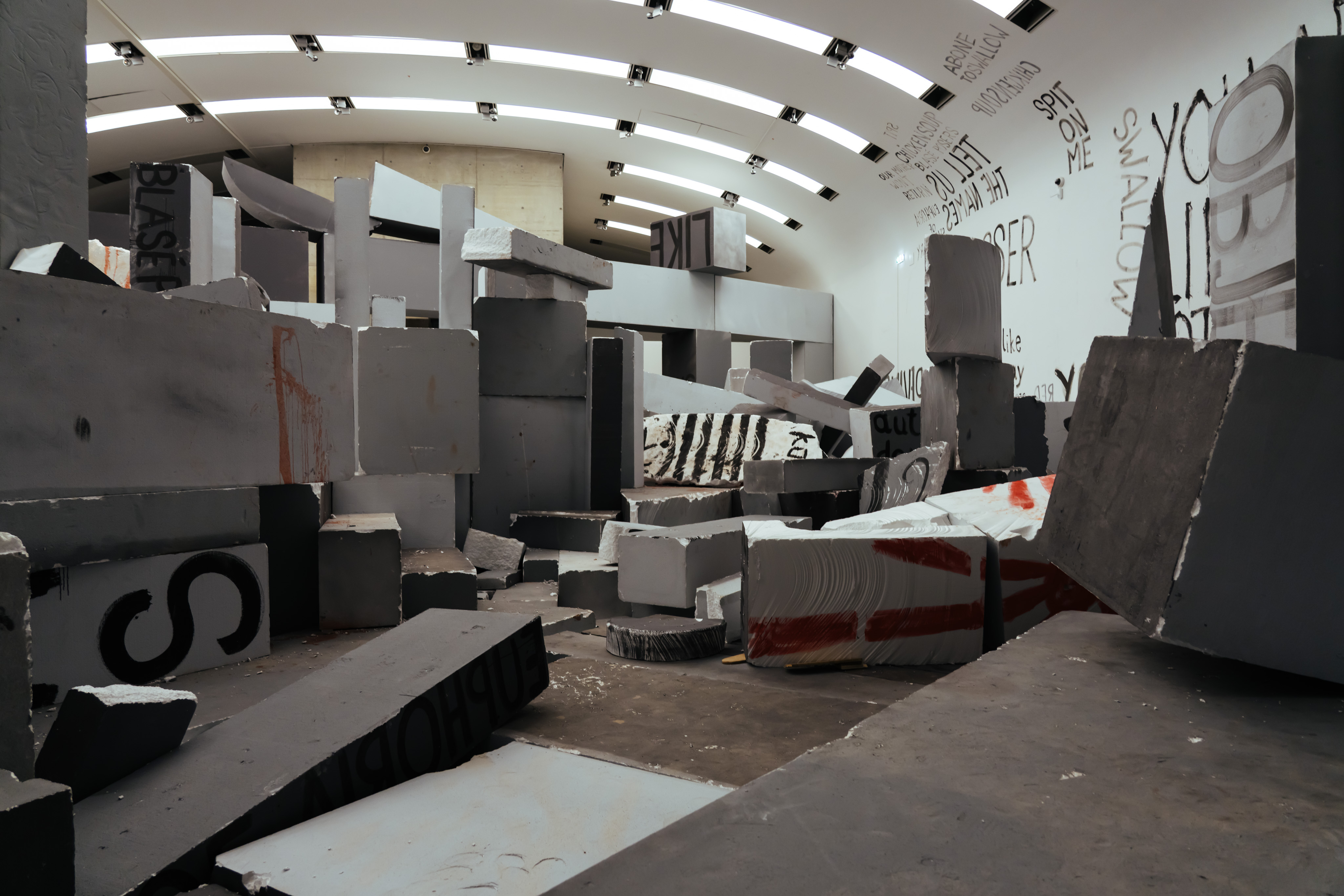 A large gallery room space with an arched ceiling is filled with large, grey styrofoam blocks. The blocks are haphazardly placed throughout the room such that it looks like ruins of a destroyed building. Many of the blocks have black or red paint on them. On the wall to the right is more graffiti reading things like "tell us the names" "spit on me" "swallow" "chicken soup blasé posers"