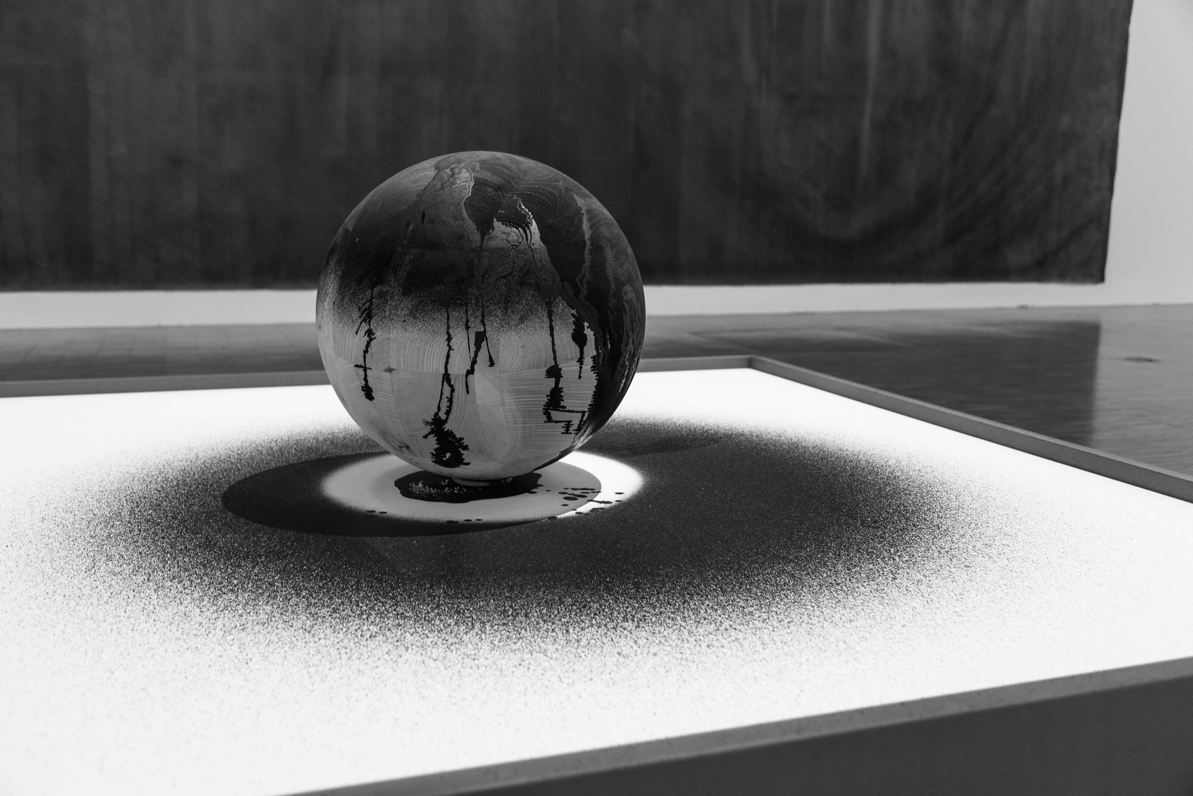 A painted sphere sits on a piece of canvas. The sphere's painting is seemingly haphazard and was done in such a way that overspray landed no parts of the canvas. There is a distinctly unpainted portion of canvas directly under the sphere and the lighting is stark such that the sphere also casts a shadow on the canvas.