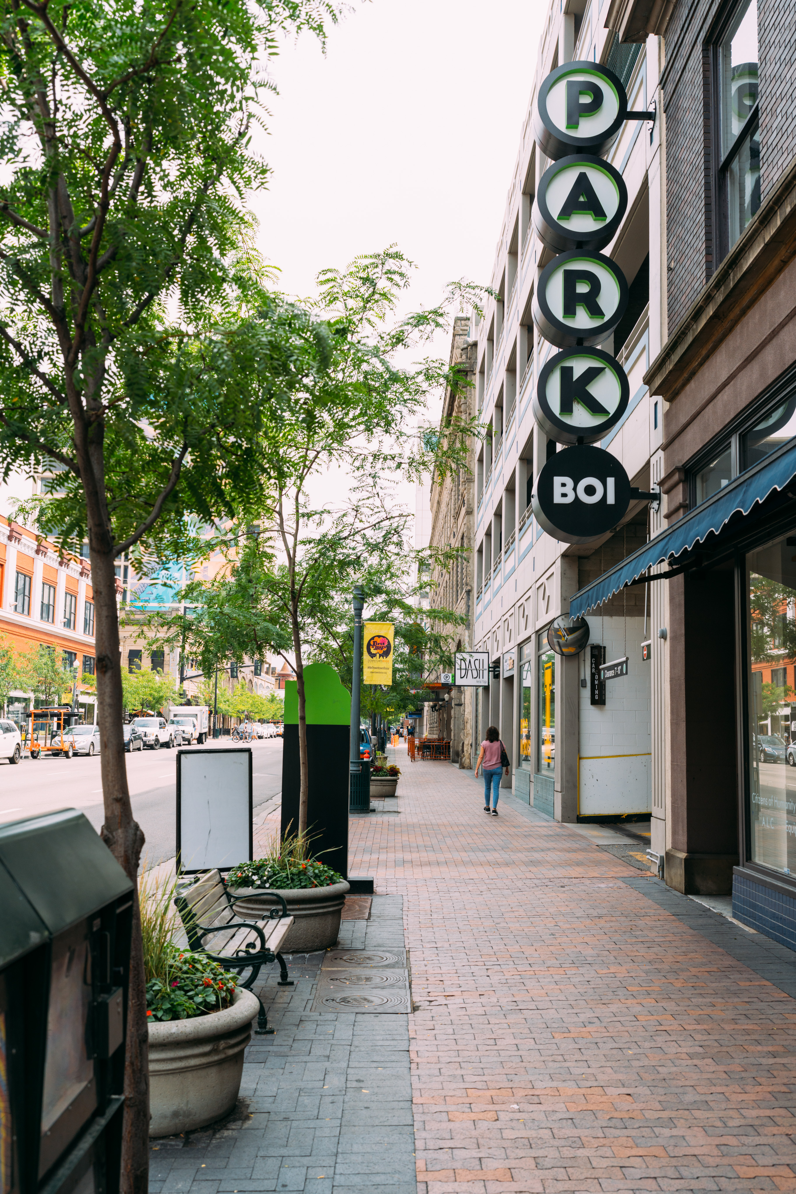 A streetscape from Boise looking down a sidewalk. There is a sign in the upper right corner that reads "Park boi". A woman walks down the sidewalk (away from the camera). Trees line the left side, with a street and cars in the background.