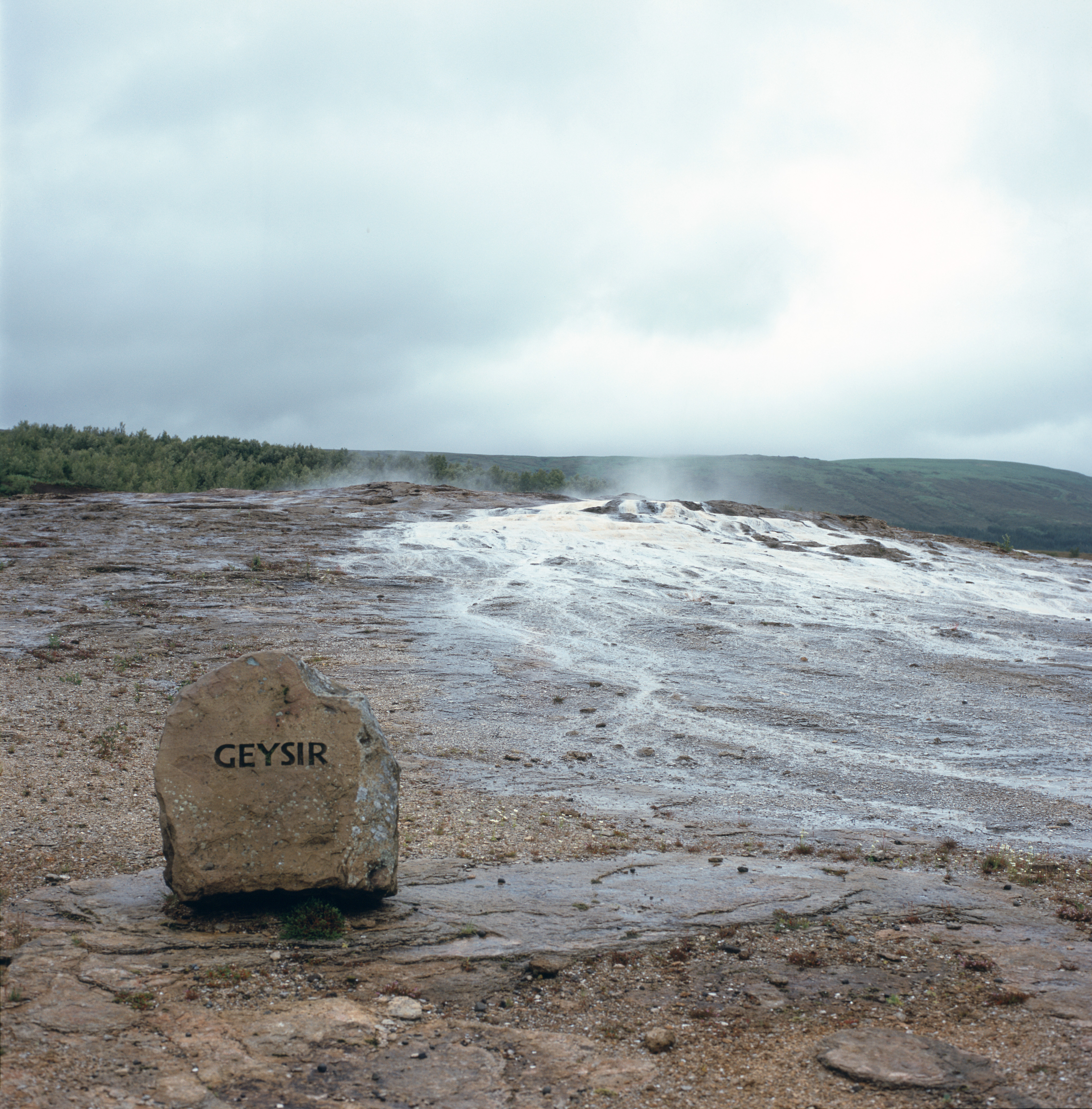 A swath of white surrounds the dormant geyser named Geysir. In the foreground is a rock with the name Geysir engraved on it.