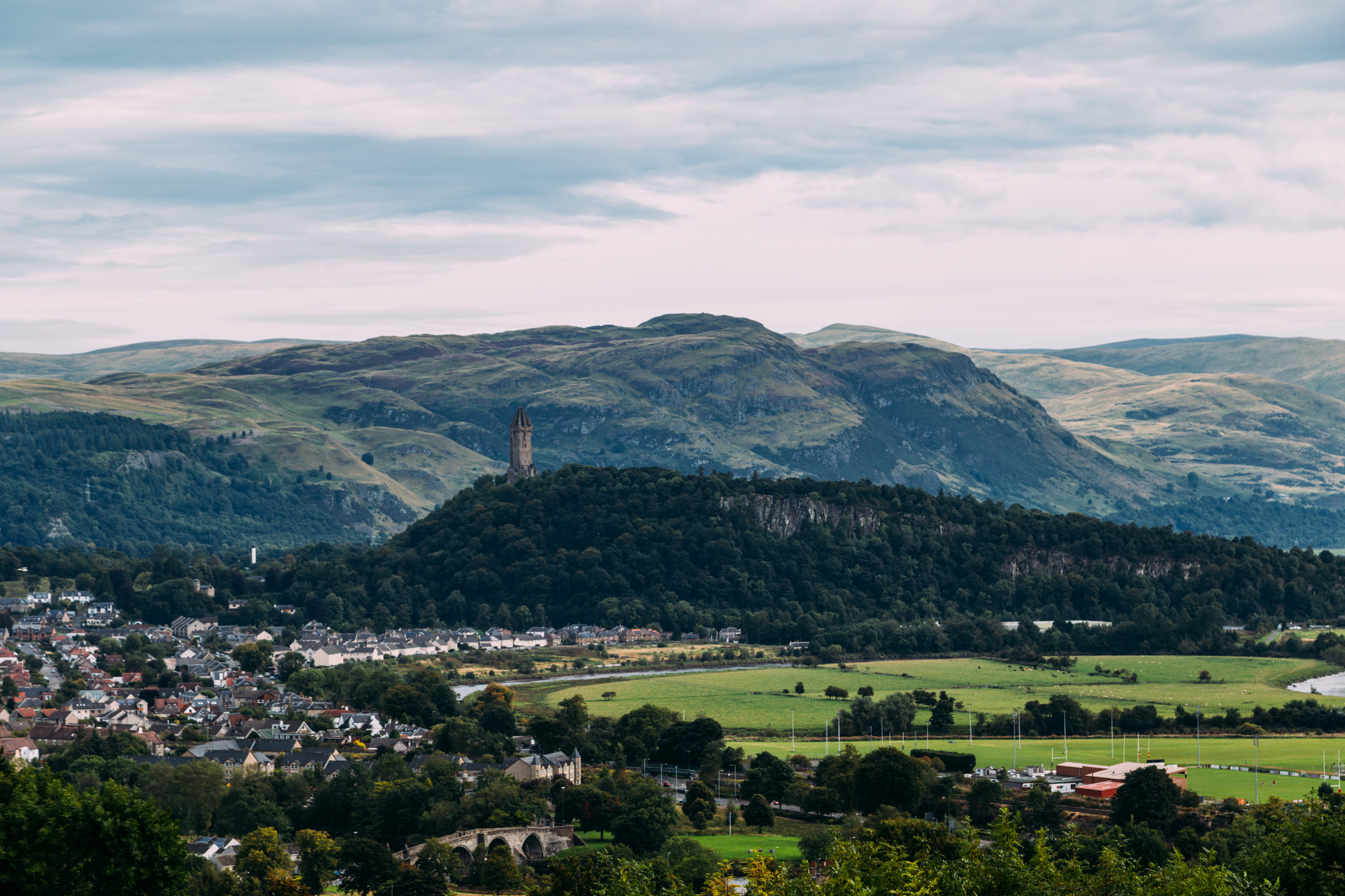 A view of the Scottish countryside looking towards the Wallace monument. On the left of the frame lay a number of buildings of a town, behind them and to the right is a large, forested hill. On top of the hill is the Wallace monument tower. In the background are rolling hills of the Scottish countryside.