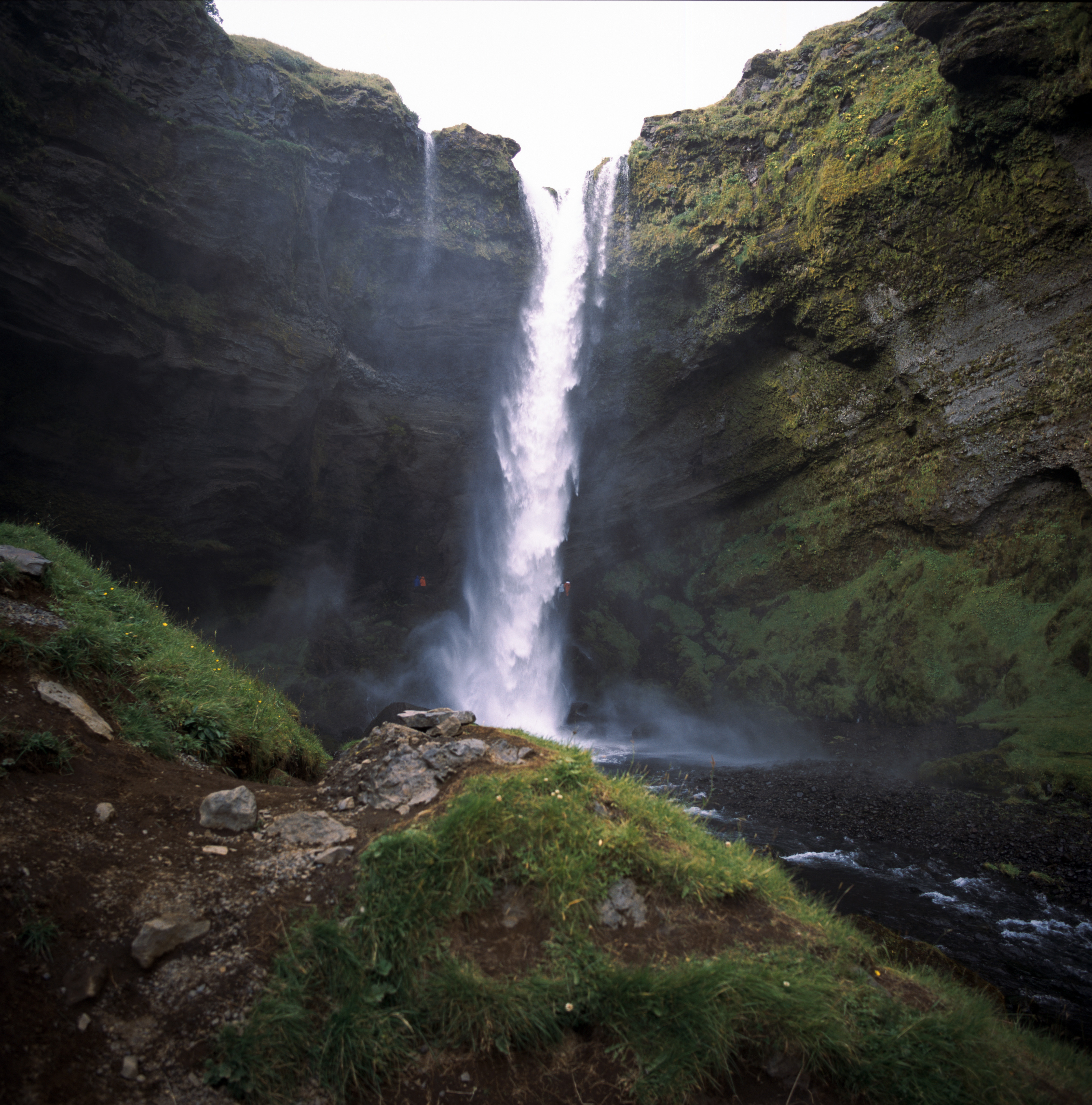 A skinny waterfall inside of a valley. The valley surrounds the entire frame. The cliff faces are black with rock and green with moss.