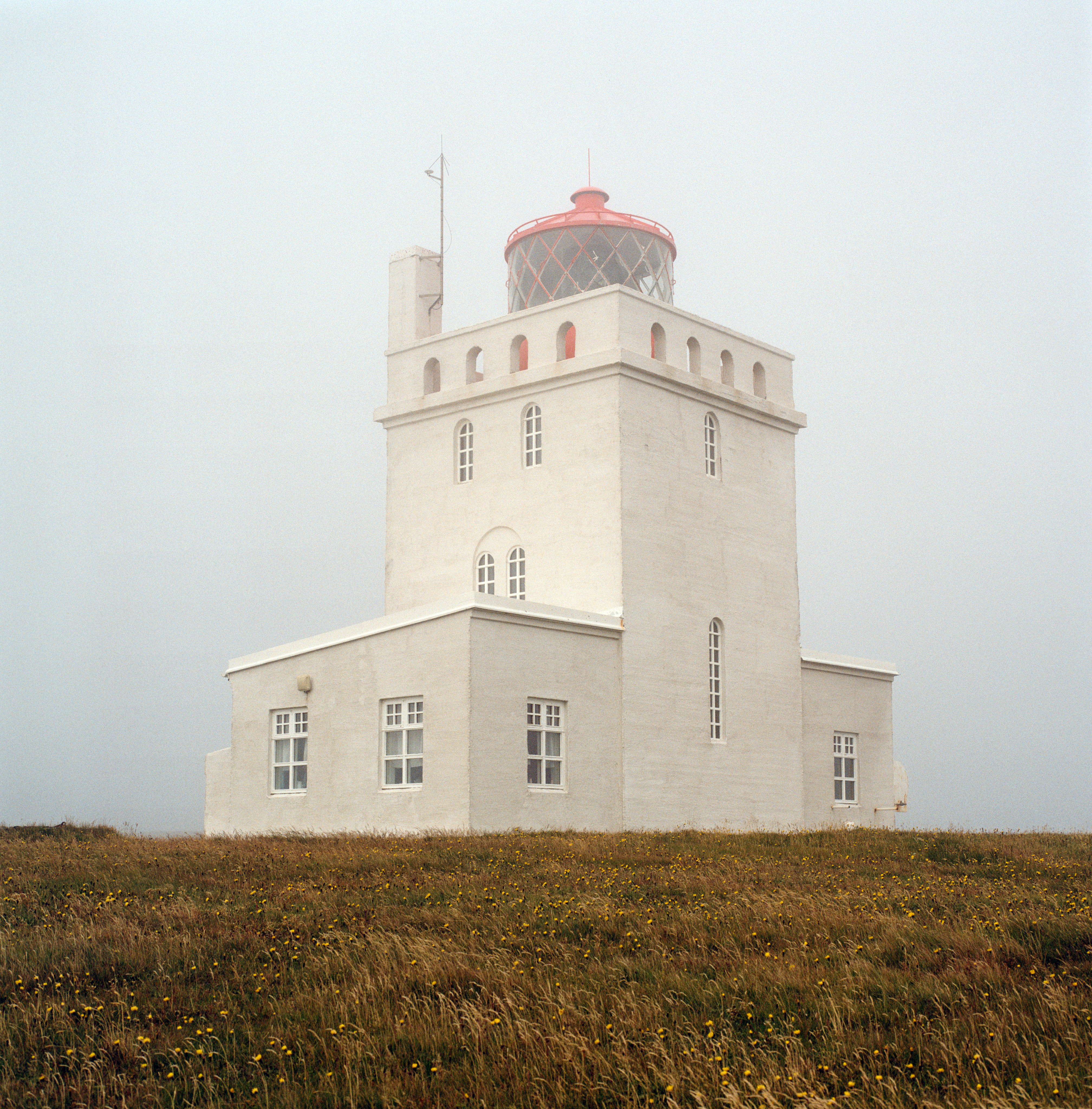 The image depicts a white, rectangular lighthouse standing tall against a foggy, overcast sky. The structure is composed of a large, square building topped by a smaller tower with a circular red dome and lattice windows, housing the light. The main body of the lighthouse features several arched windows that are symmetrically placed. The walls are painted in a clean, pale hue that contrasts with the red of the dome. In the foreground, there is a field of grass scattered with small yellow flowers, adding color to the composition. The overall atmosphere is calm and serene, with the fog providing a sense of isolation and tranquility.